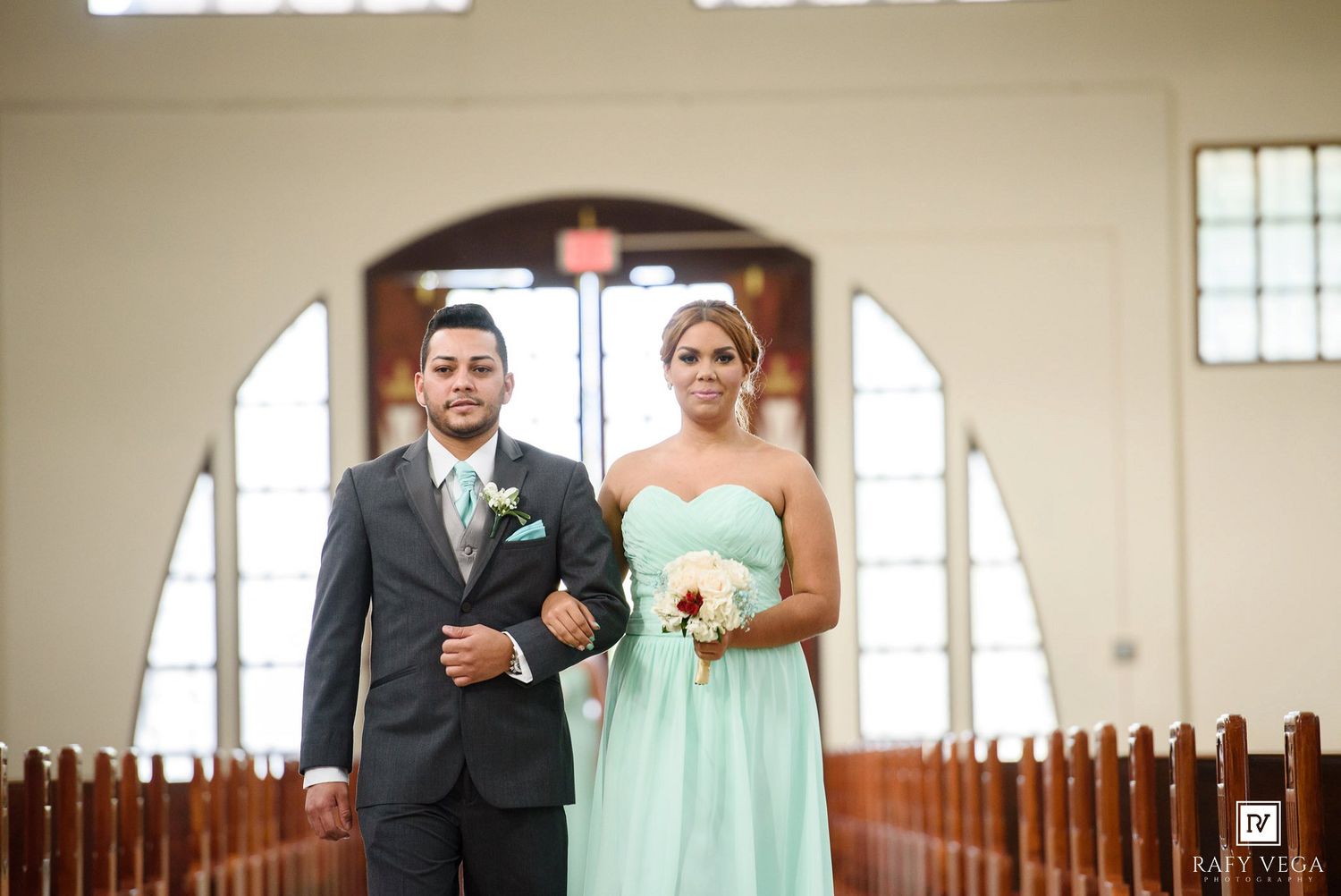 A groomsman in a gray suit escorts a bridesmaid in a mint green gown down a church aisle during a wedding ceremony.