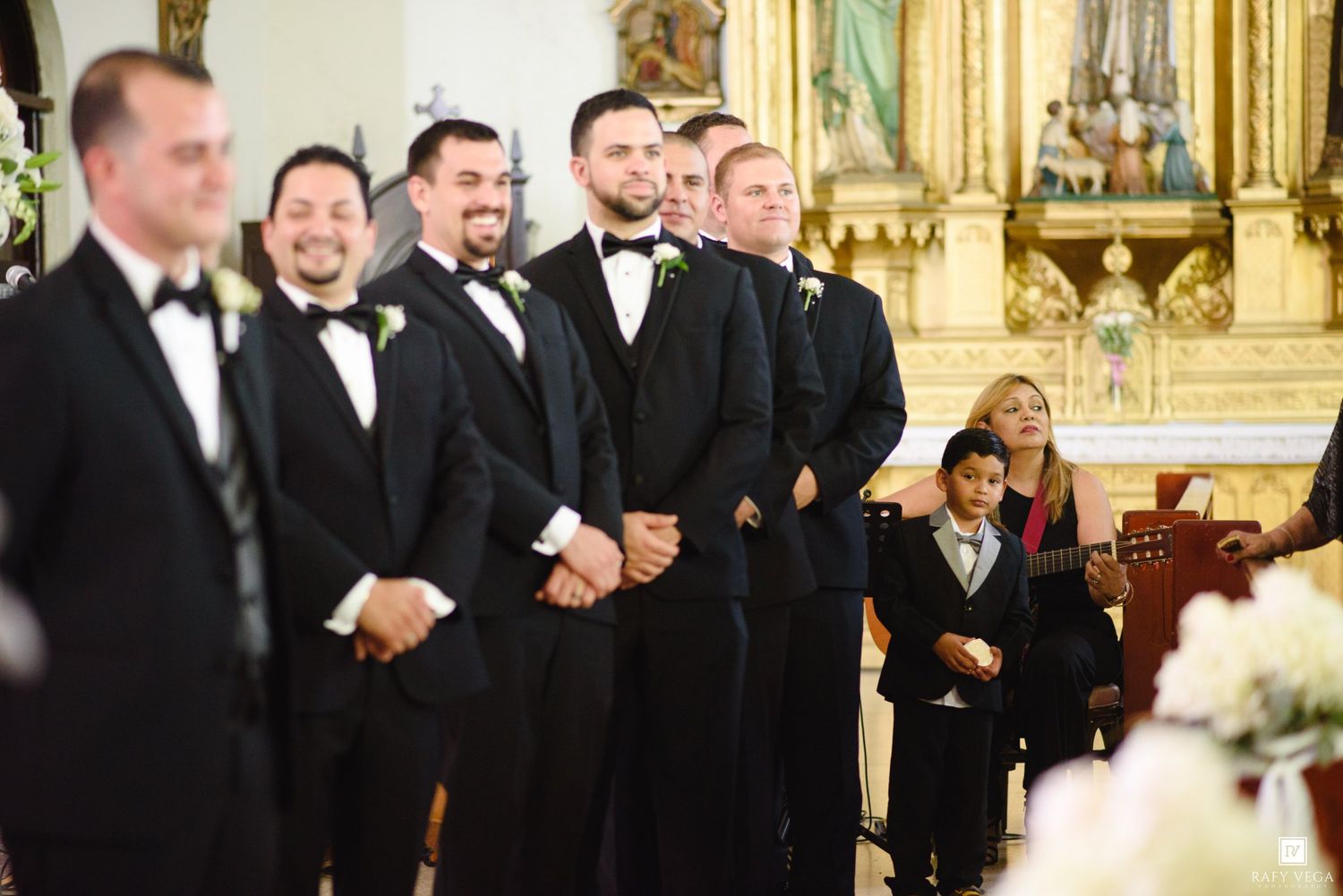 Groomsmen in black tuxedos stand at the altar during a wedding ceremony inside an ornate church.