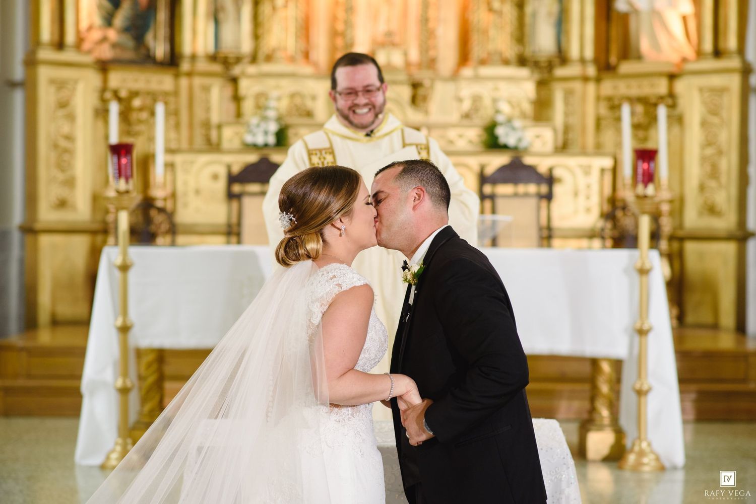 A bride and groom share their first kiss at the altar during a church wedding ceremony with a smiling priest.