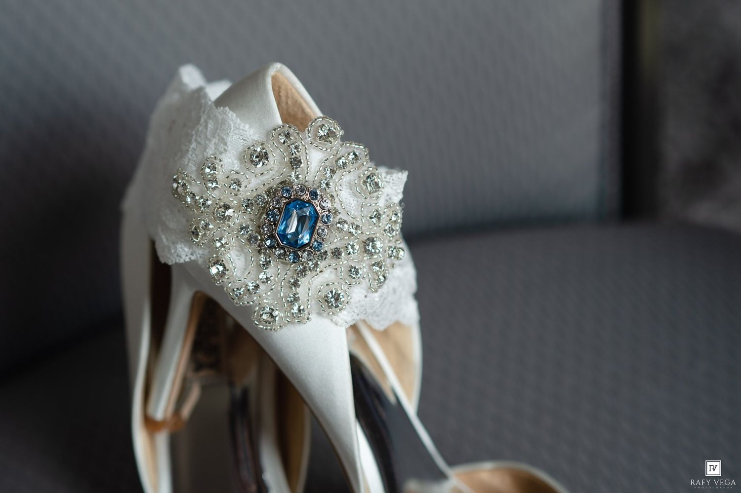 White bridal heels adorned with sparkling rhinestone brooch featuring a blue sapphire center stone on a gray background.