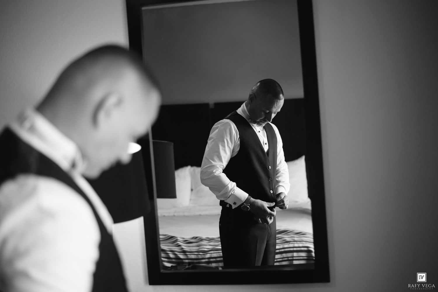 Groom adjusting vest in mirror reflection, black and white wedding photo.