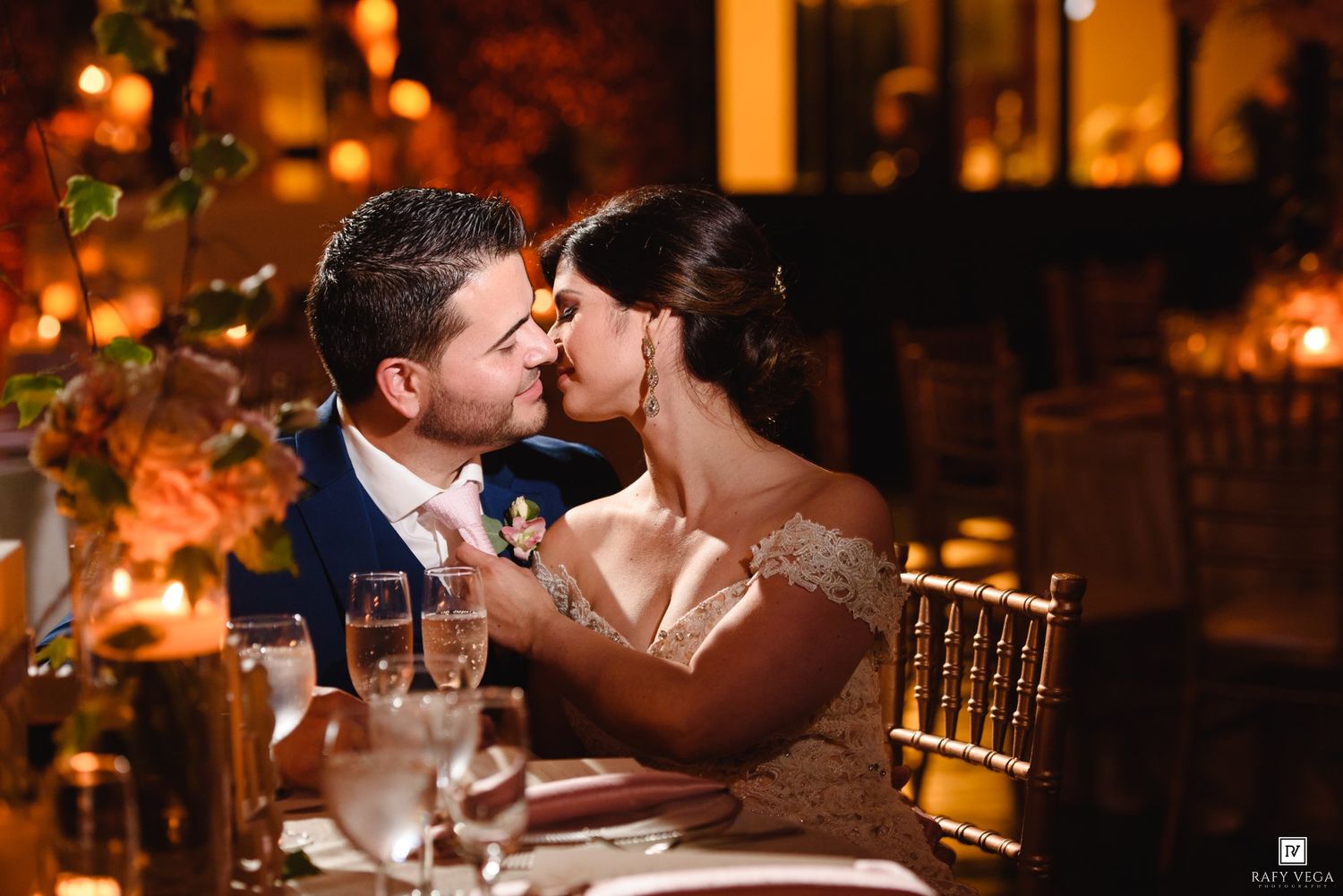 Bride and groom share a romantic kiss at candlelit wedding reception table with warm golden lighting.
