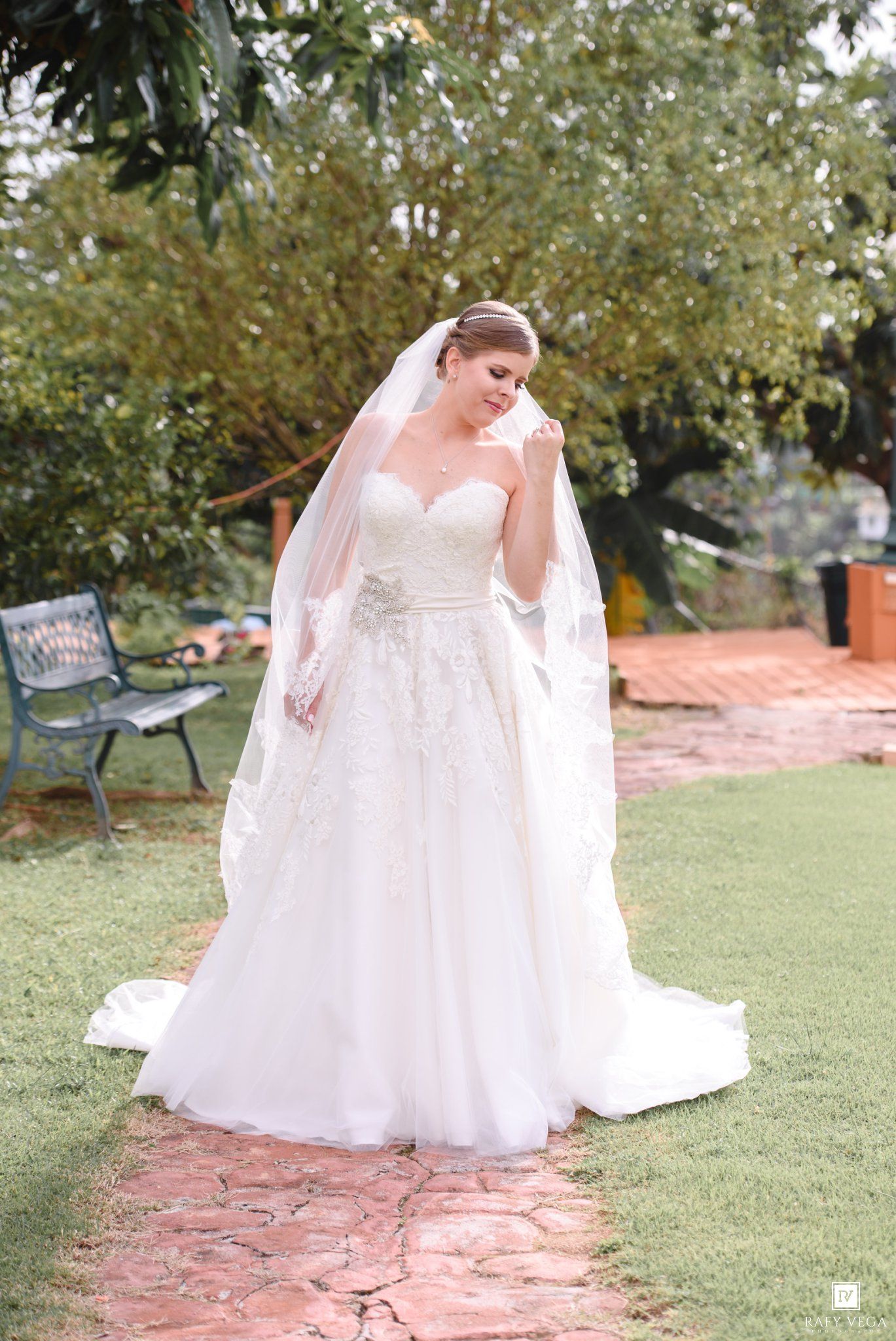 Bride in a strapless lace ball gown with long veil stands on brick path in garden setting.