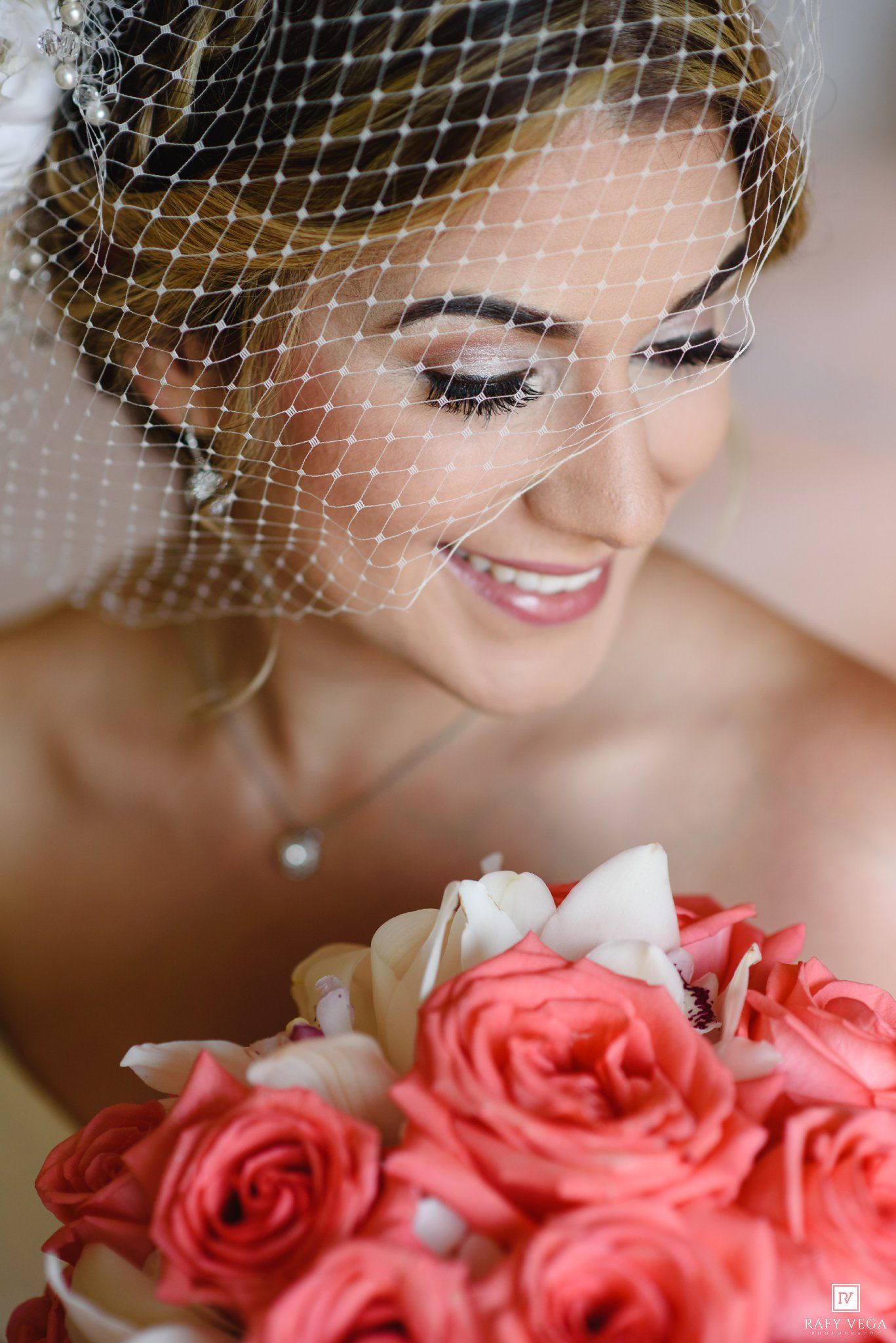 Smiling bride wearing a birdcage veil holds a bouquet of coral and cream roses on her wedding day.
