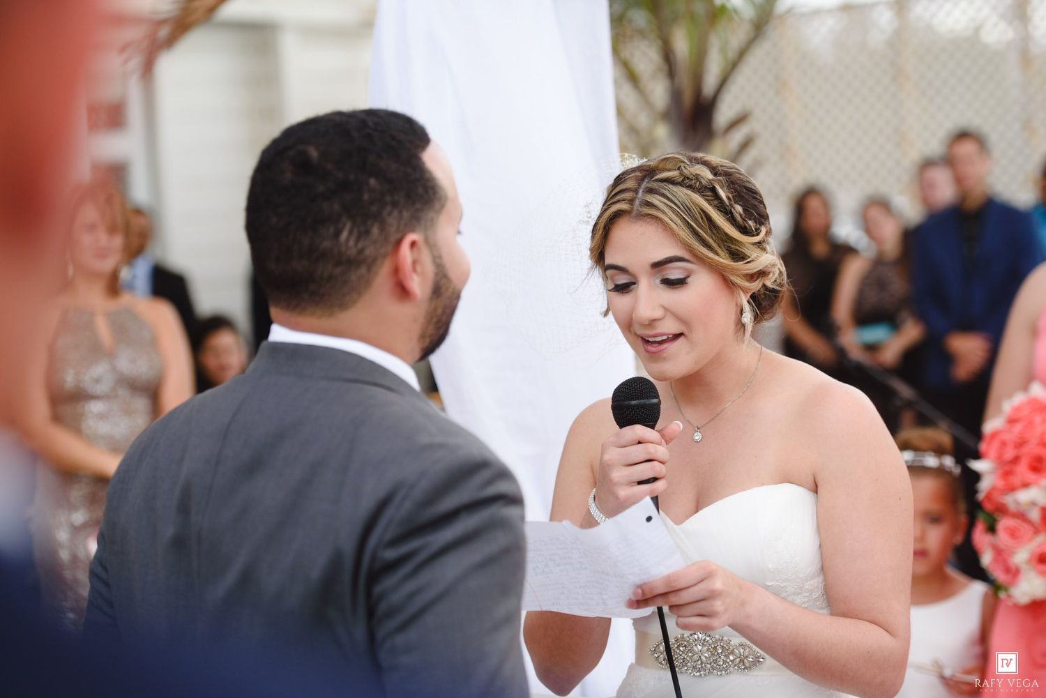 Bride in strapless gown reads vows into microphone at outdoor wedding ceremony.