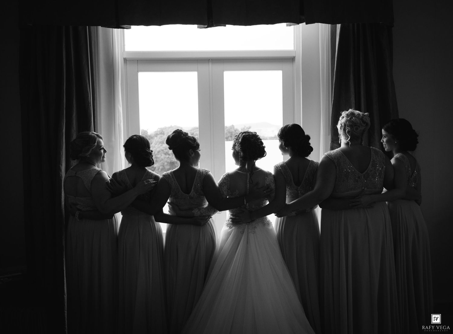 Black and white photo of bride and bridesmaids standing together by a bright window in elegant gowns.