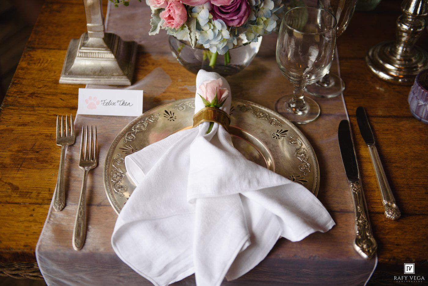 Elegant wedding table setting with gold charger plate, white napkin, and floral centerpiece on wood table.