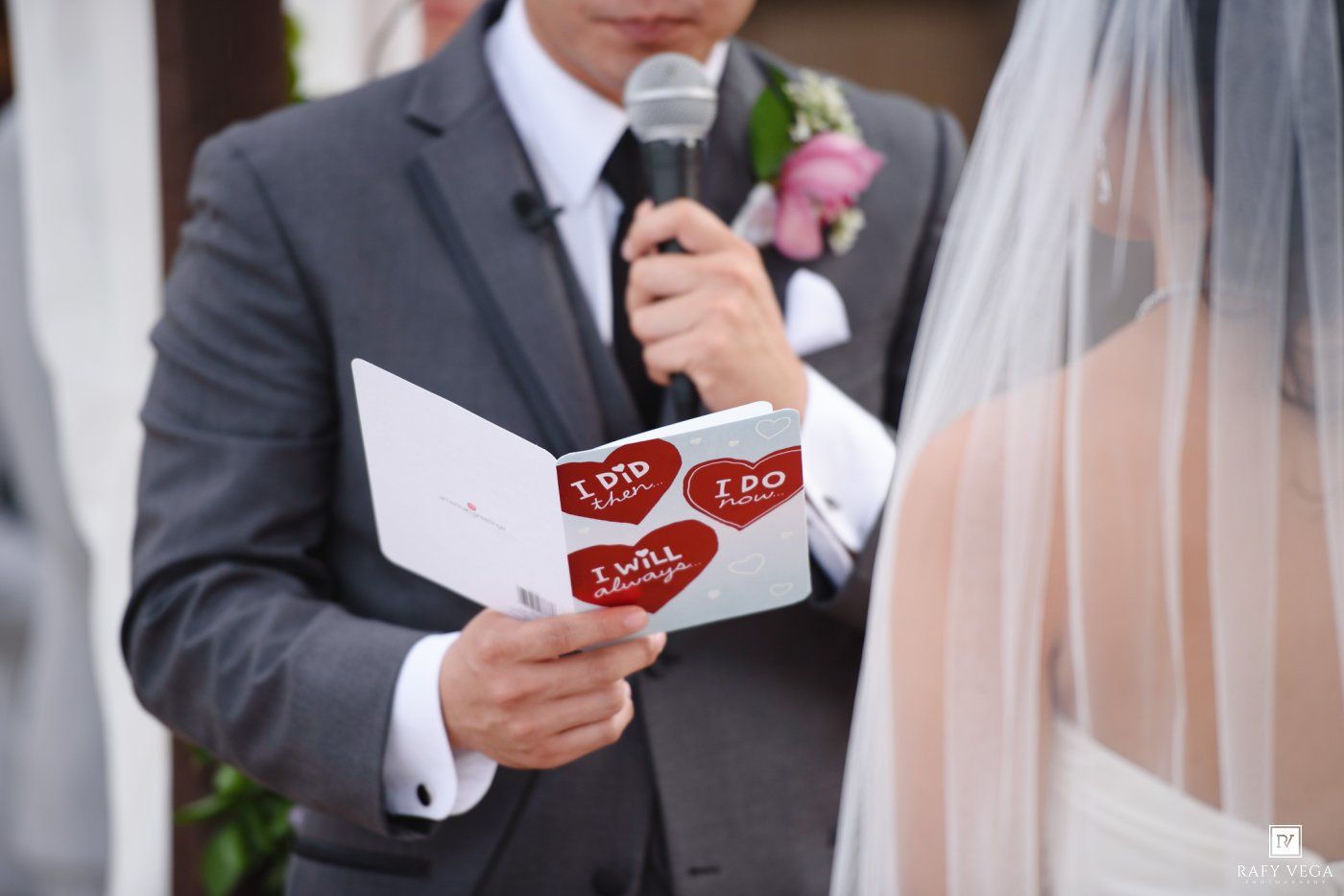 Groom reading wedding vows from a booklet with red heart designs during an outdoor wedding ceremony.