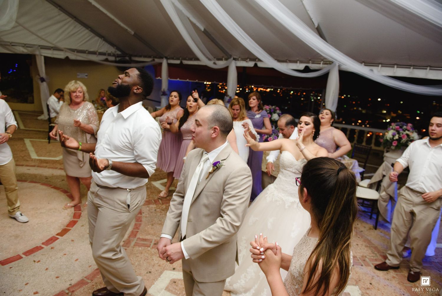 Guests dancing joyfully at an outdoor wedding reception under a white tent at night, with the bride visible in the background.