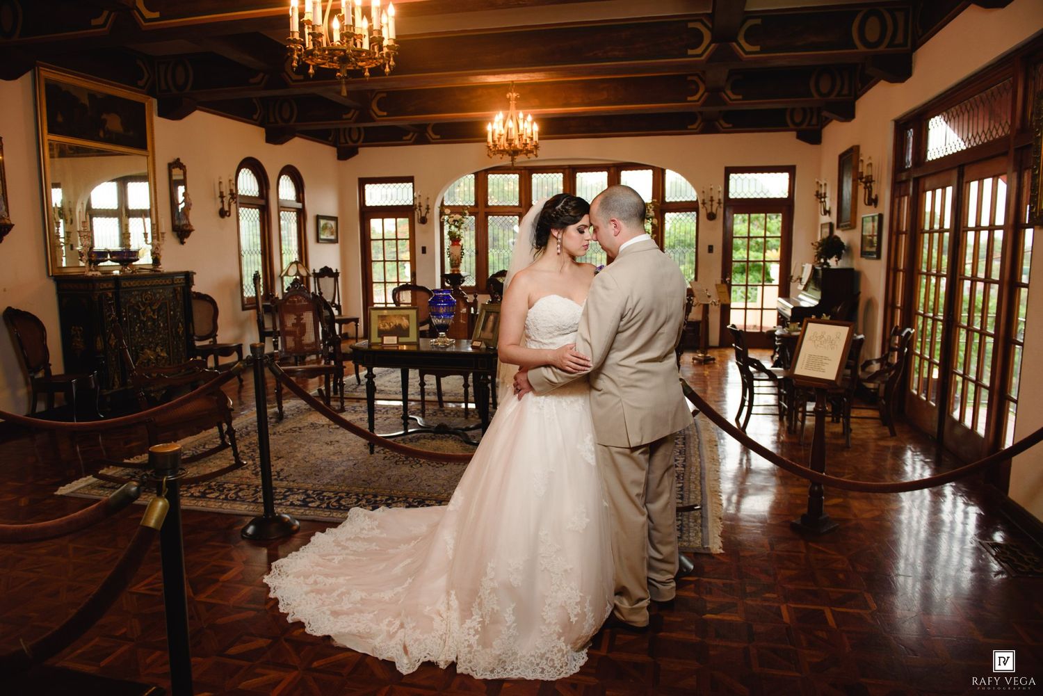 Bride and groom share a romantic first dance in an elegant wood-paneled ballroom with chandeliers and arched windows.