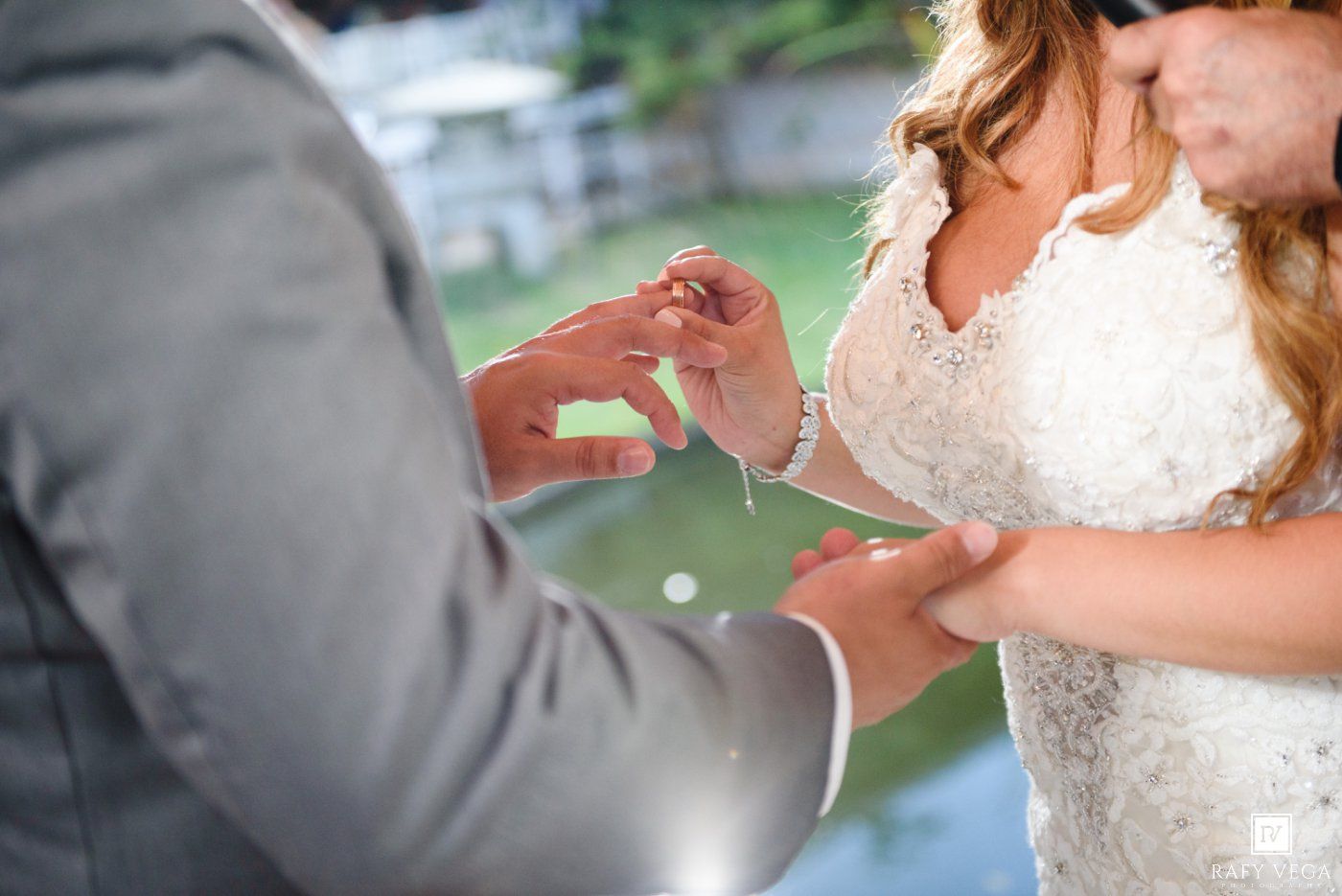 Groom in gray suit placing wedding ring on brides finger during outdoor ceremony, bride wearing lace white gown.