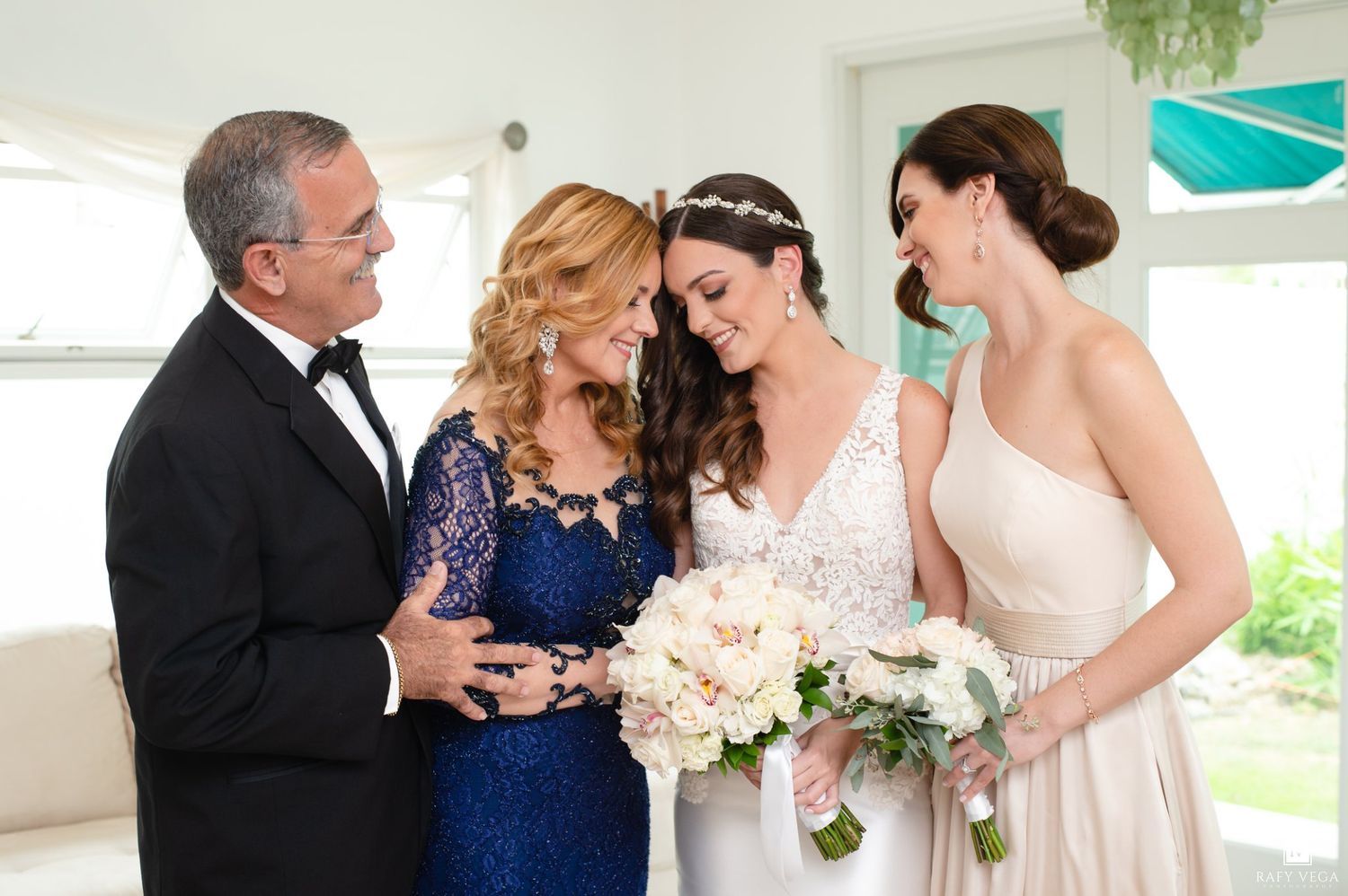 Bride in lace gown shares tender moment with family including mother in blue lace dress.