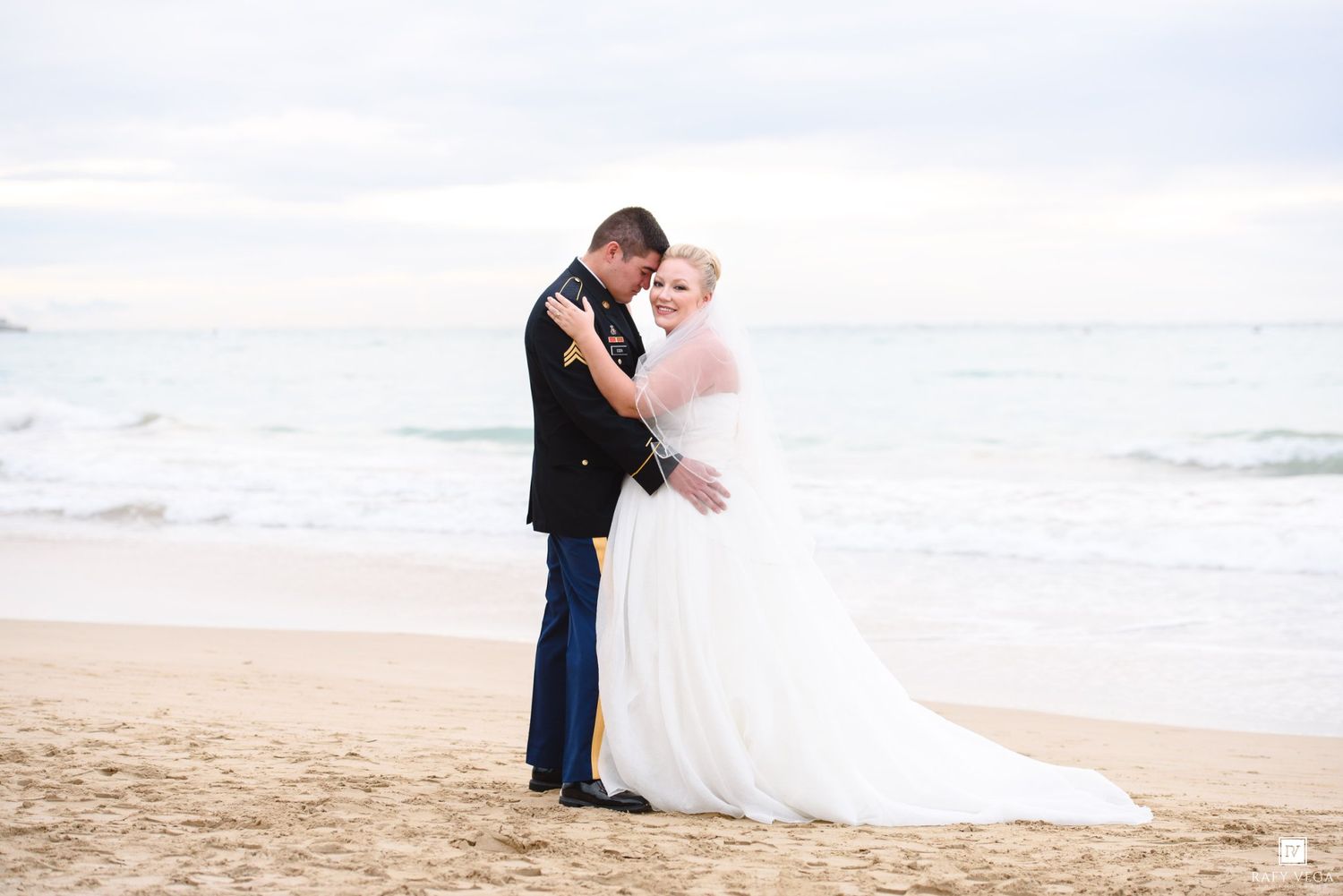 Bride and groom embracing on a sandy beach, with ocean waves in the background on their wedding day.