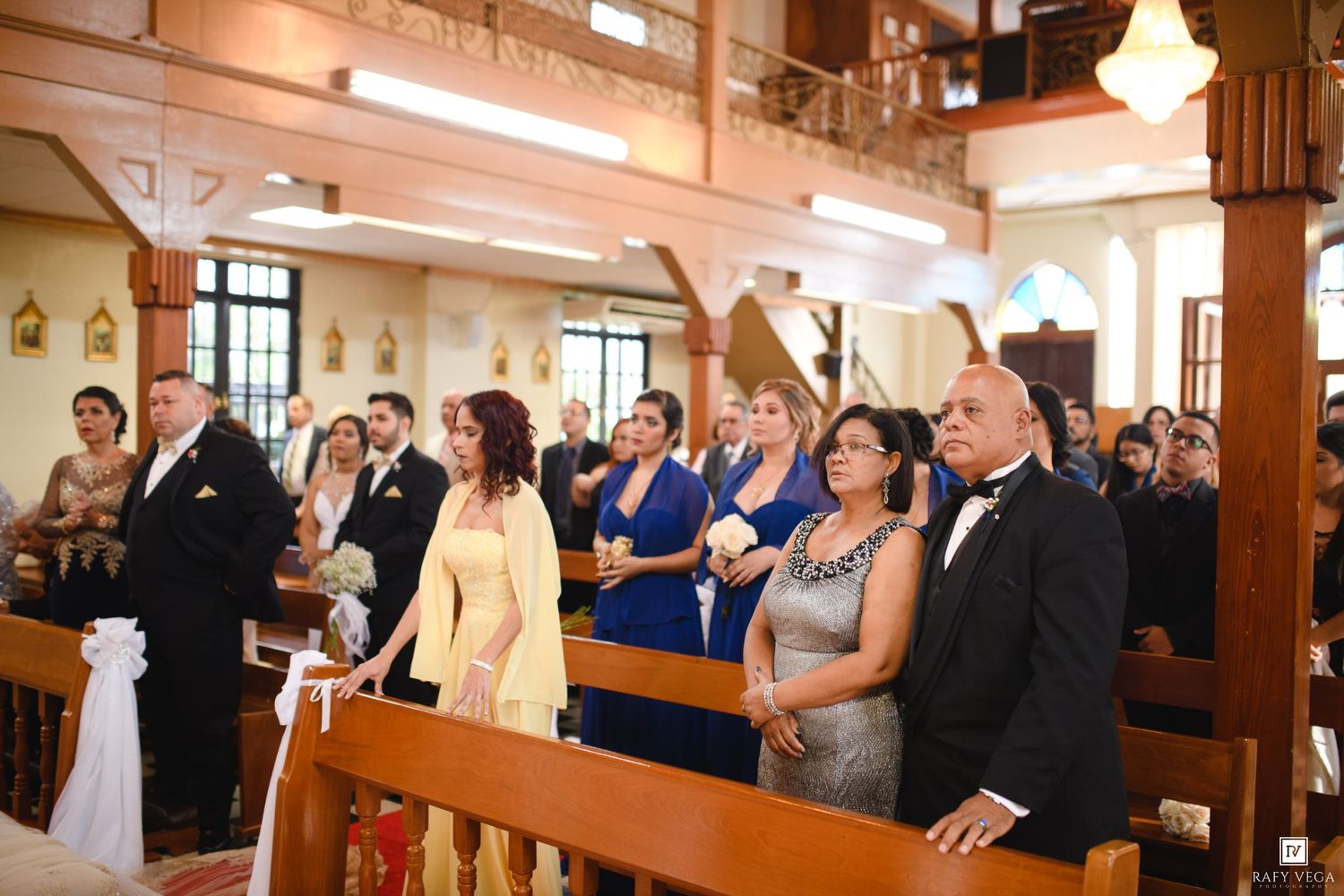 Guests standing in church pews during a wedding ceremony, women in blue and yellow dresses.