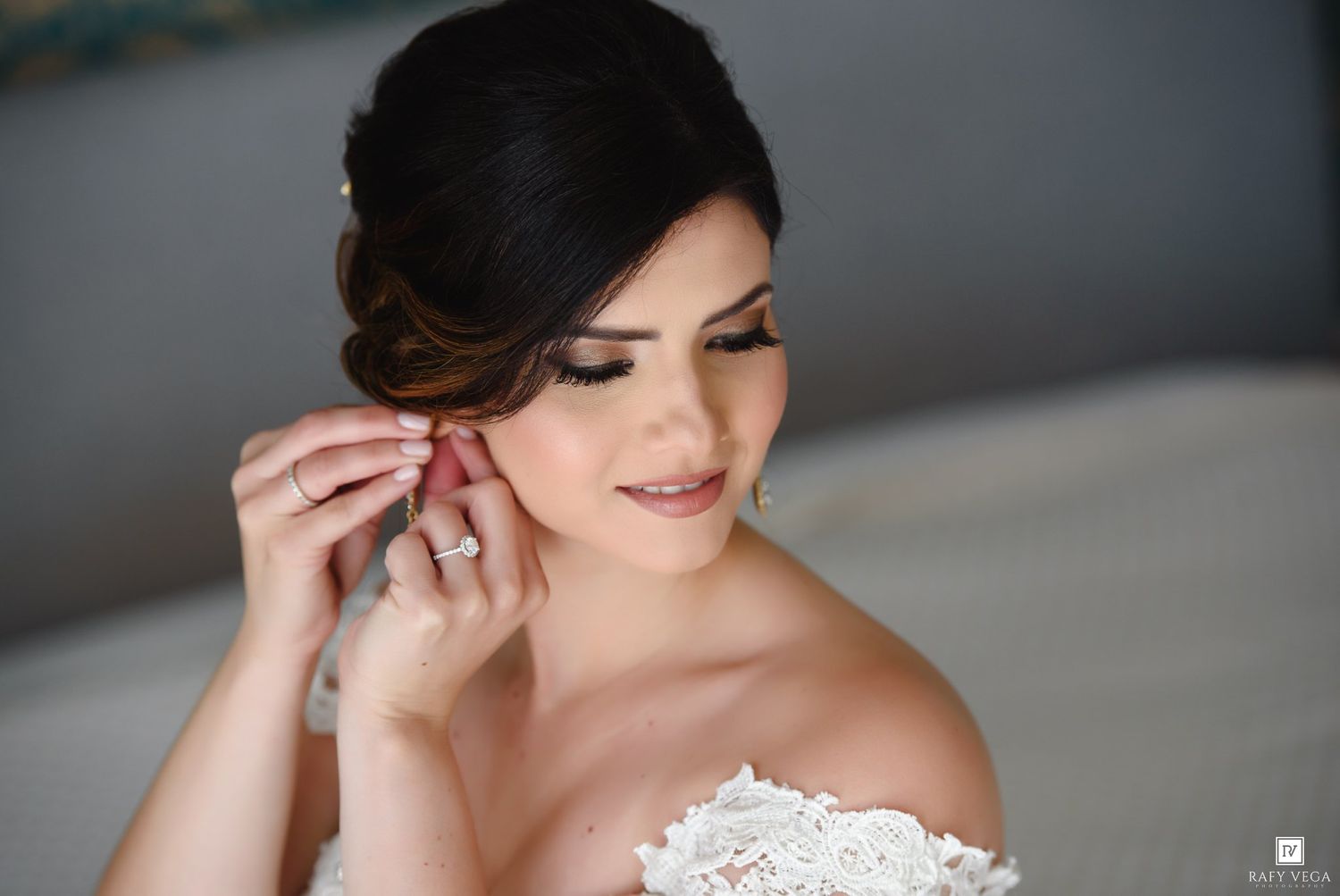Bride in strapless lace gown putting on earring, wearing diamond ring, with elegant updo hairstyle and natural makeup.