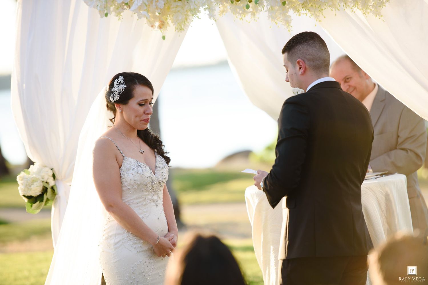 Bride and groom exchange vows at an outdoor waterfront wedding ceremony under a white draped arch.