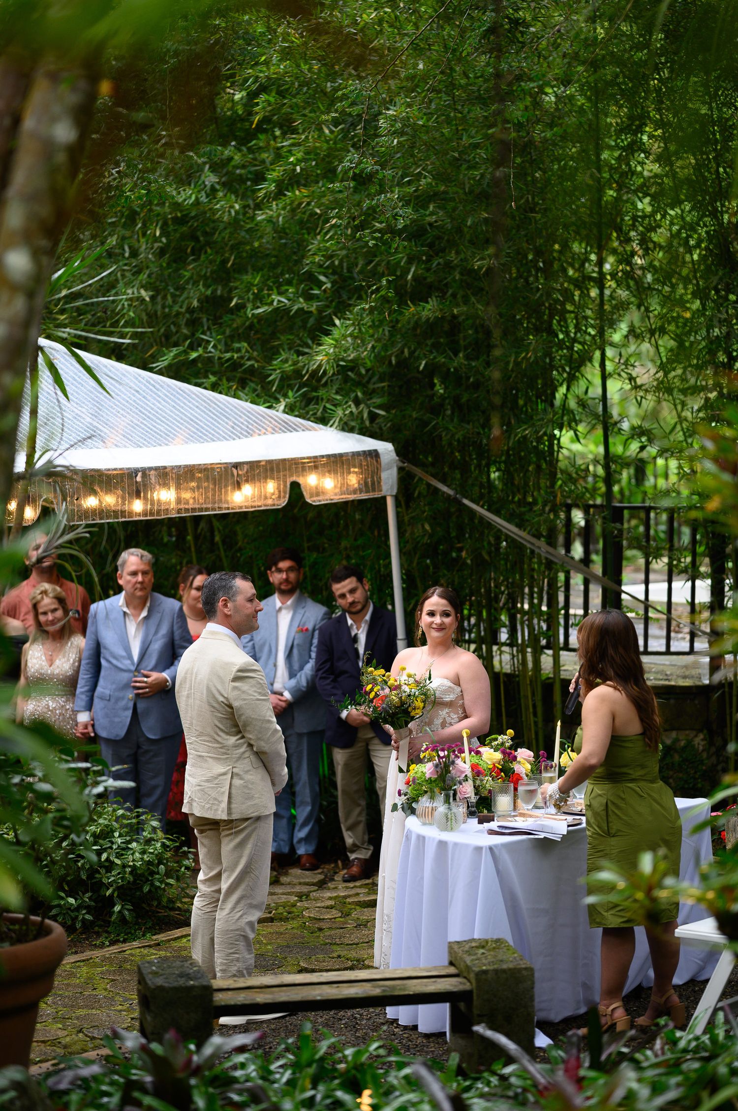 Outdoor garden wedding reception with guests gathered under a lit tent surrounded by lush bamboo trees and floral arrangements.
