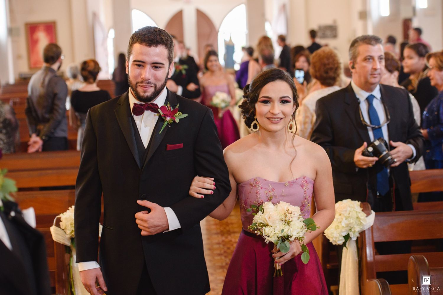 A groomsman and bridesmaid in burgundy dress walk down church aisle during wedding ceremony.
