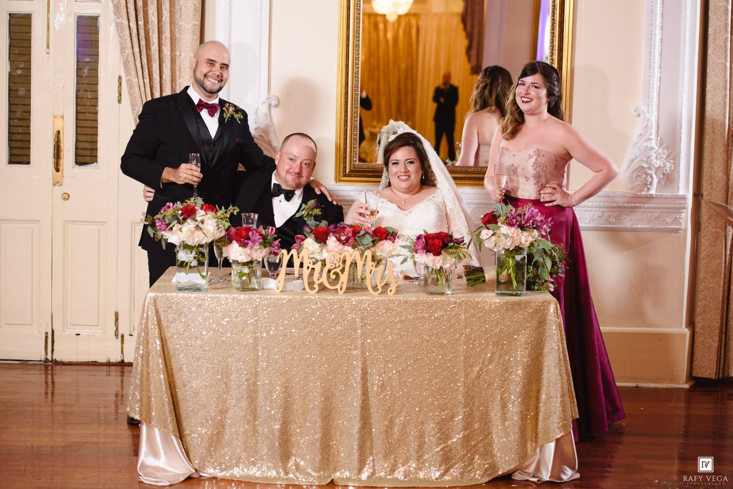 Wedding reception sweetheart table with bride and groom seated, flanked by guests in formal attire, gold sequin tablecloth.