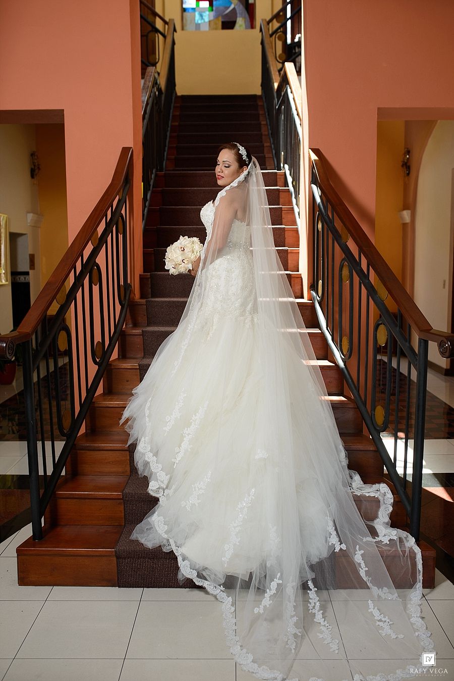 Bride in elegant white lace wedding gown with long veil holding bouquet on grand staircase.
