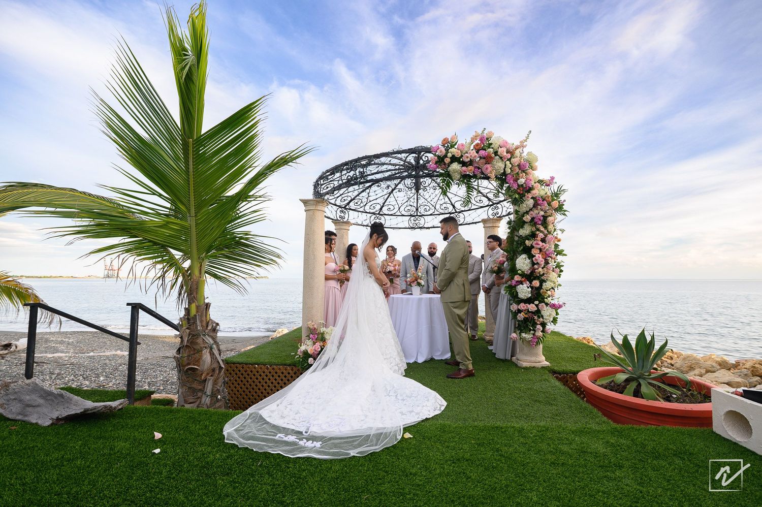 Bride in a long white gown and groom exchange vows under a floral arch on a green lawn overlooking the ocean at sunset.