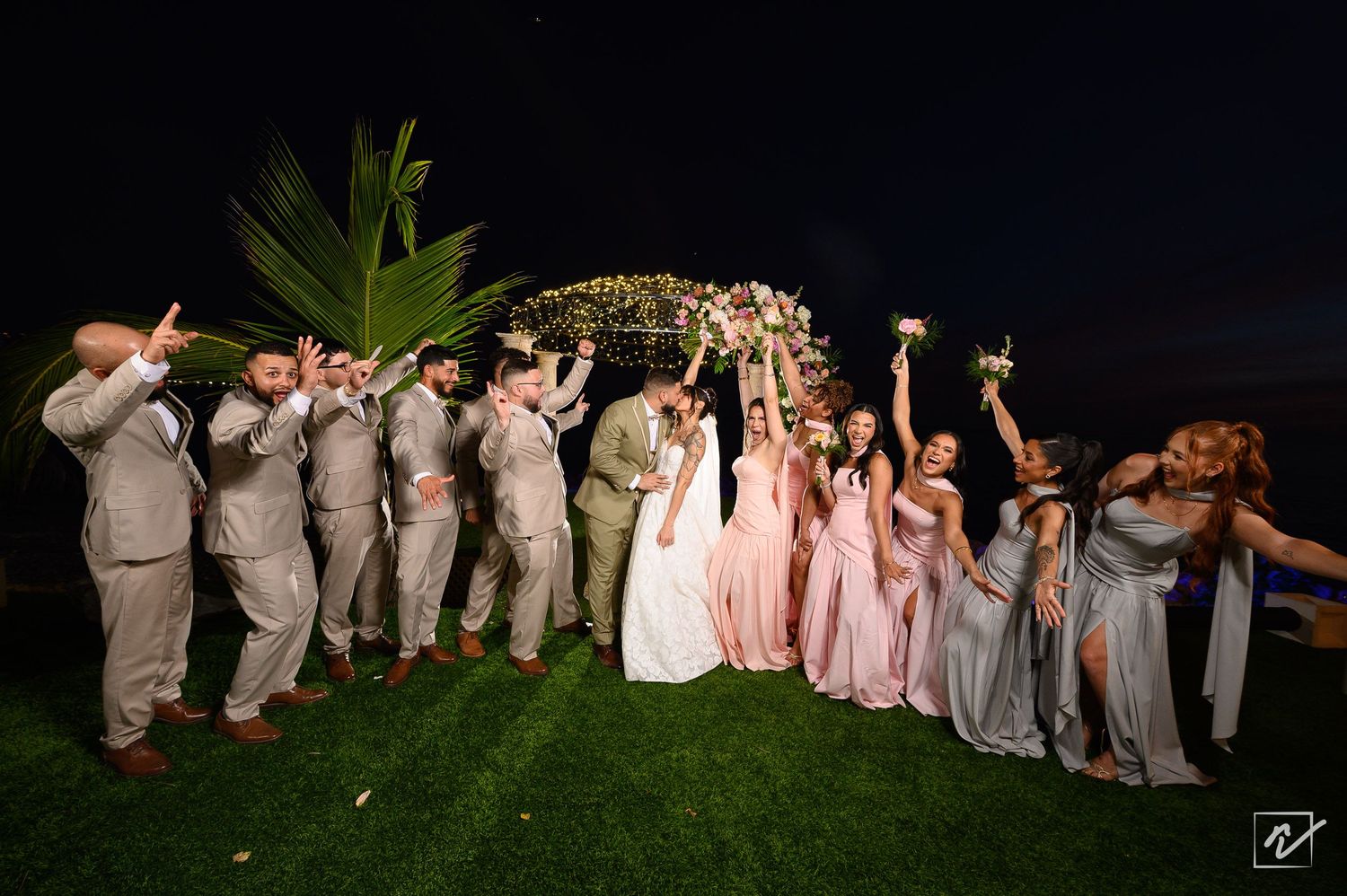 Bridal party in pink and gray dresses and tan suits cheers with raised bouquets beside the bride and groom at a night garden wedding.