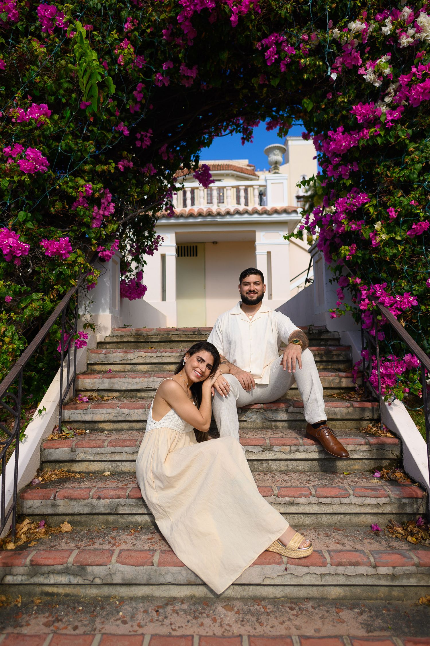 Couple in white outfits sitting on brick steps framed by vibrant pink bougainvillea with a white building behind.