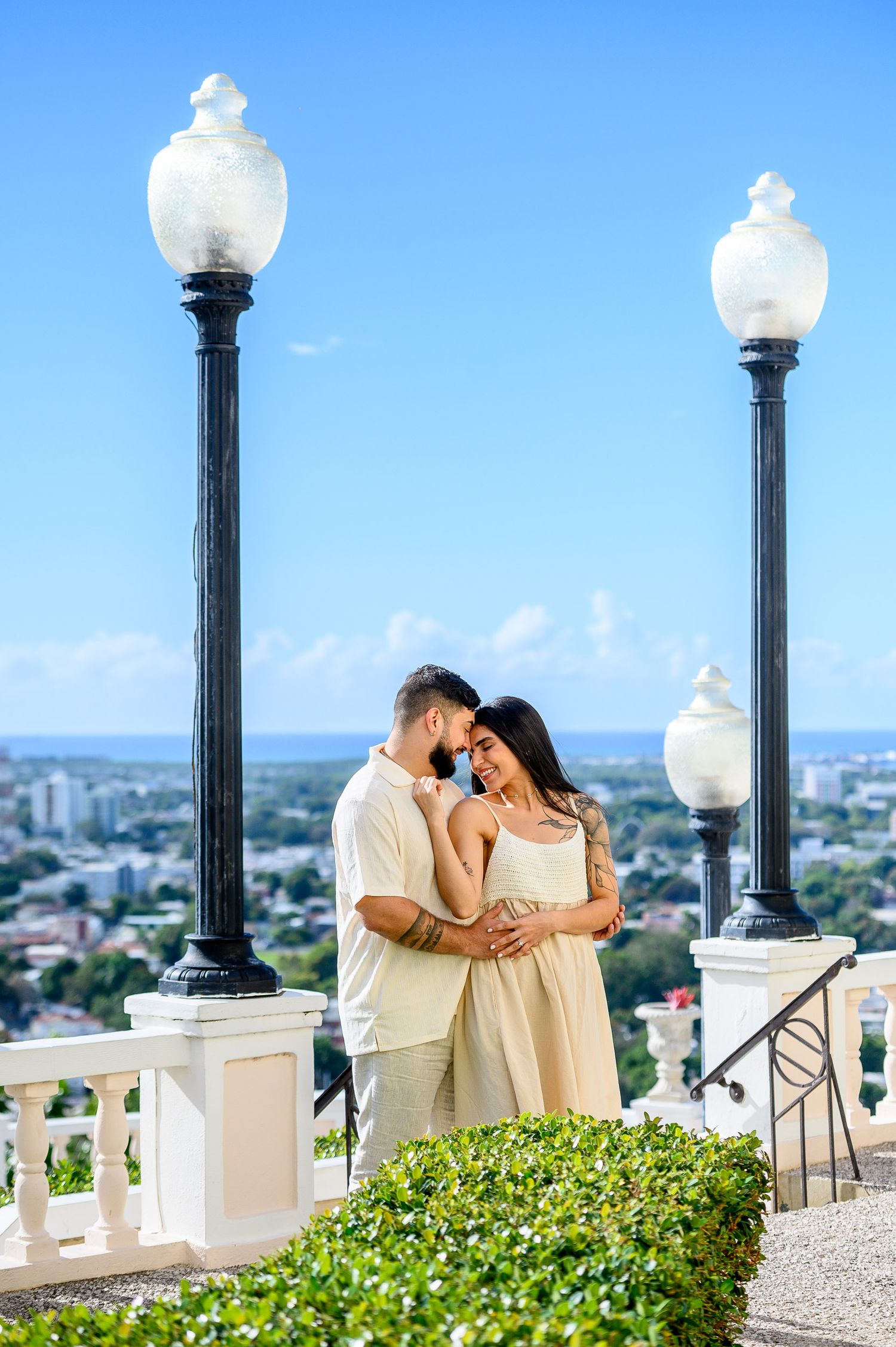 Romantic couple embracing on a scenic overlook with ornate street lamps and a city skyline under a bright blue sky.
