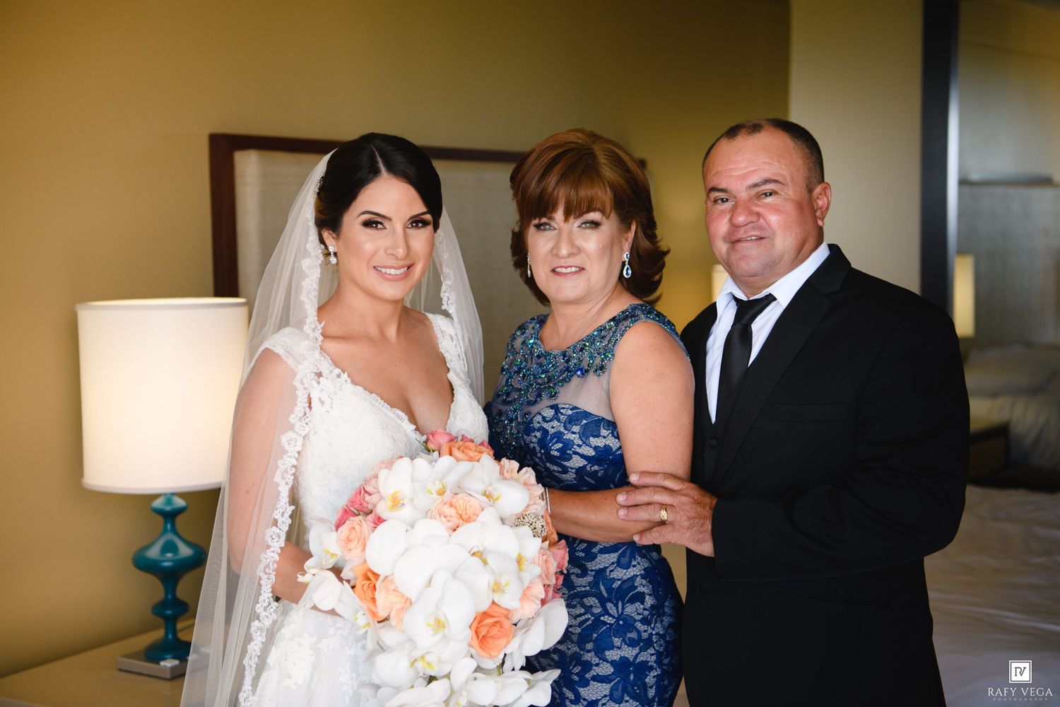 Bride in white gown holds bouquet flanked by parents in blue dress and black suit indoors.