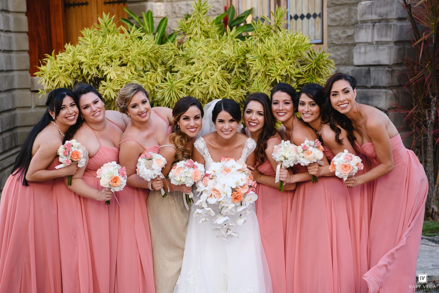 Bride in white gown poses with eight bridesmaids in coral pink dresses holding white and peach bouquets outdoors.