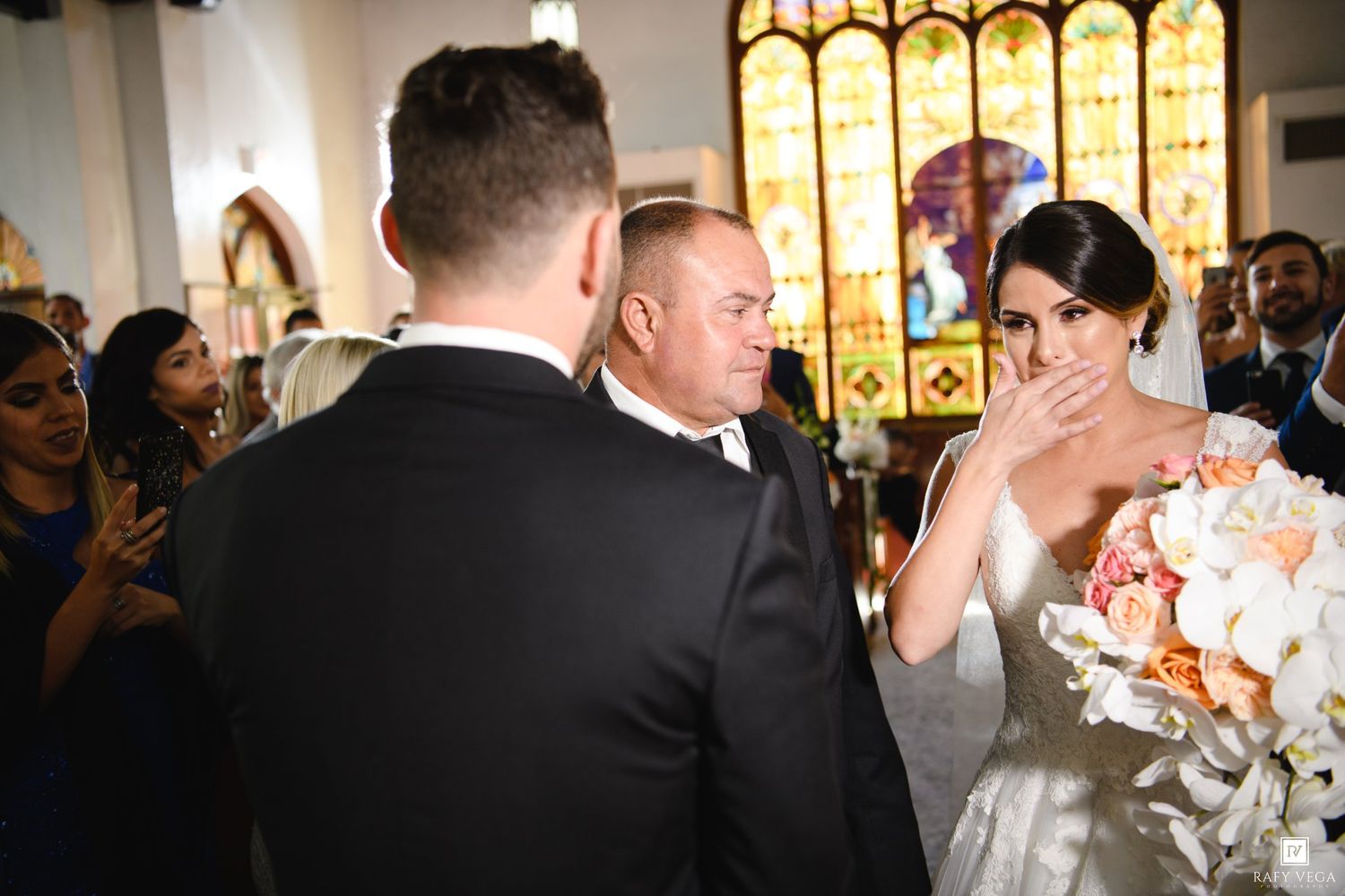 Emotional bride covering her mouth while meeting groom at church altar during wedding ceremony with stained glass windows.