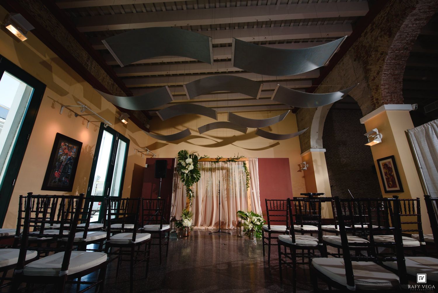 Elegant wedding ceremony setup in a historic venue with dark wood chairs, floral arch backdrop, and ornate coffered ceiling.