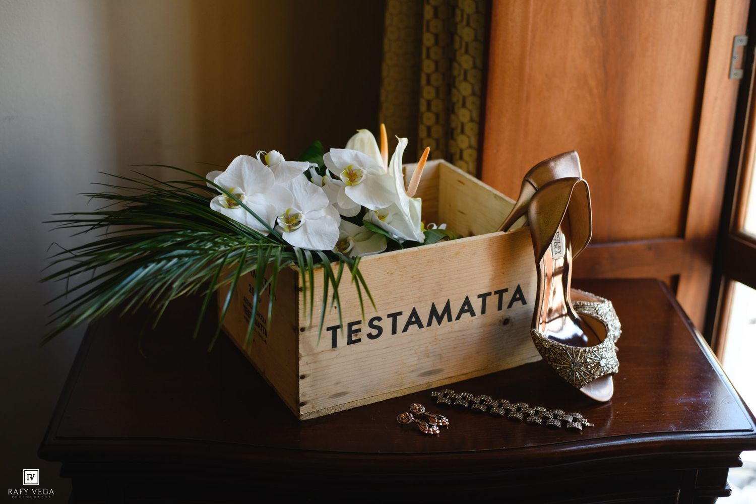 Wooden Testamatta wine crate with white orchids, green foliage, gold heels, and jewelry on a dark wood table.