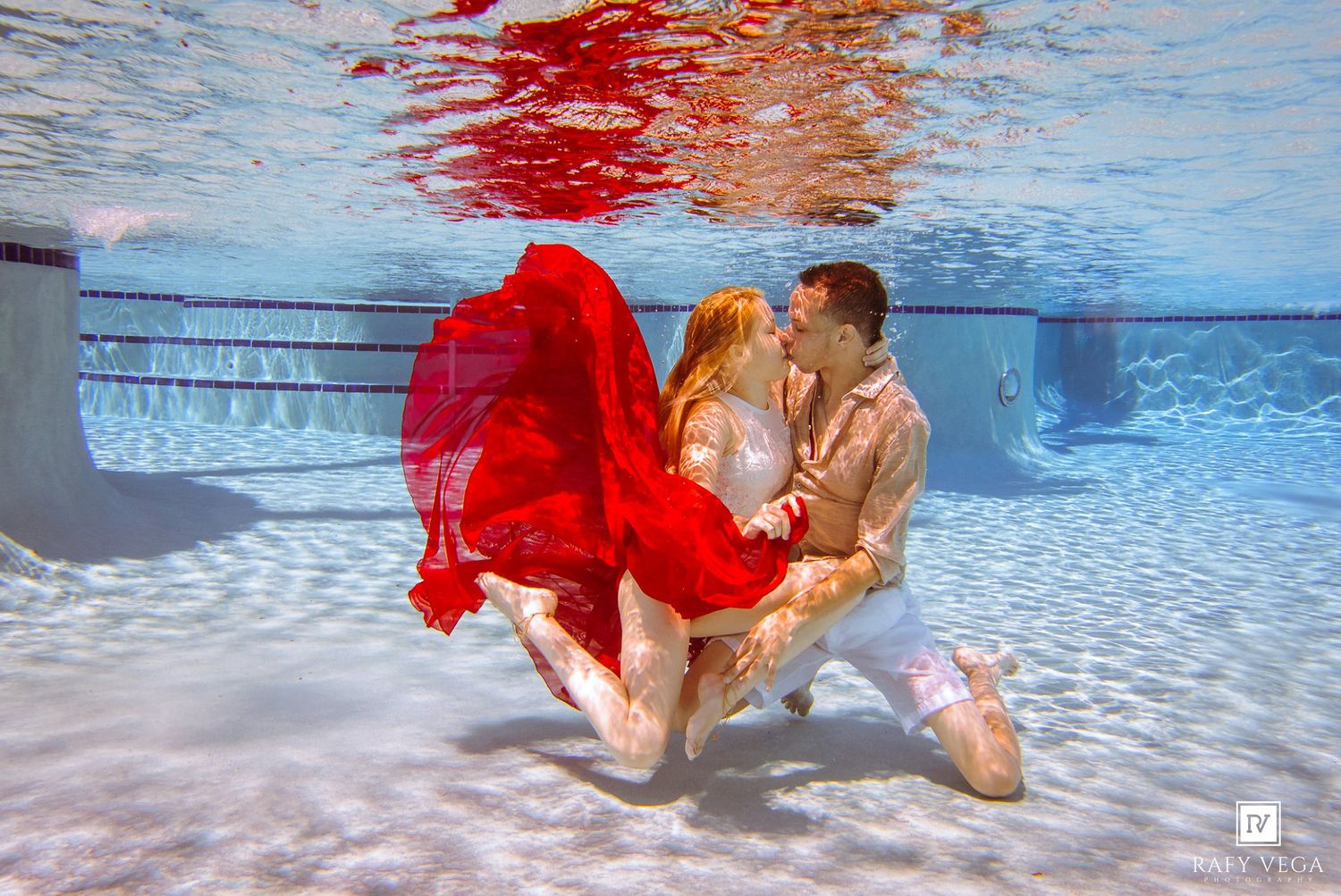 Couple kissing underwater in a pool, woman wearing a flowing red dress, romantic underwater photography.