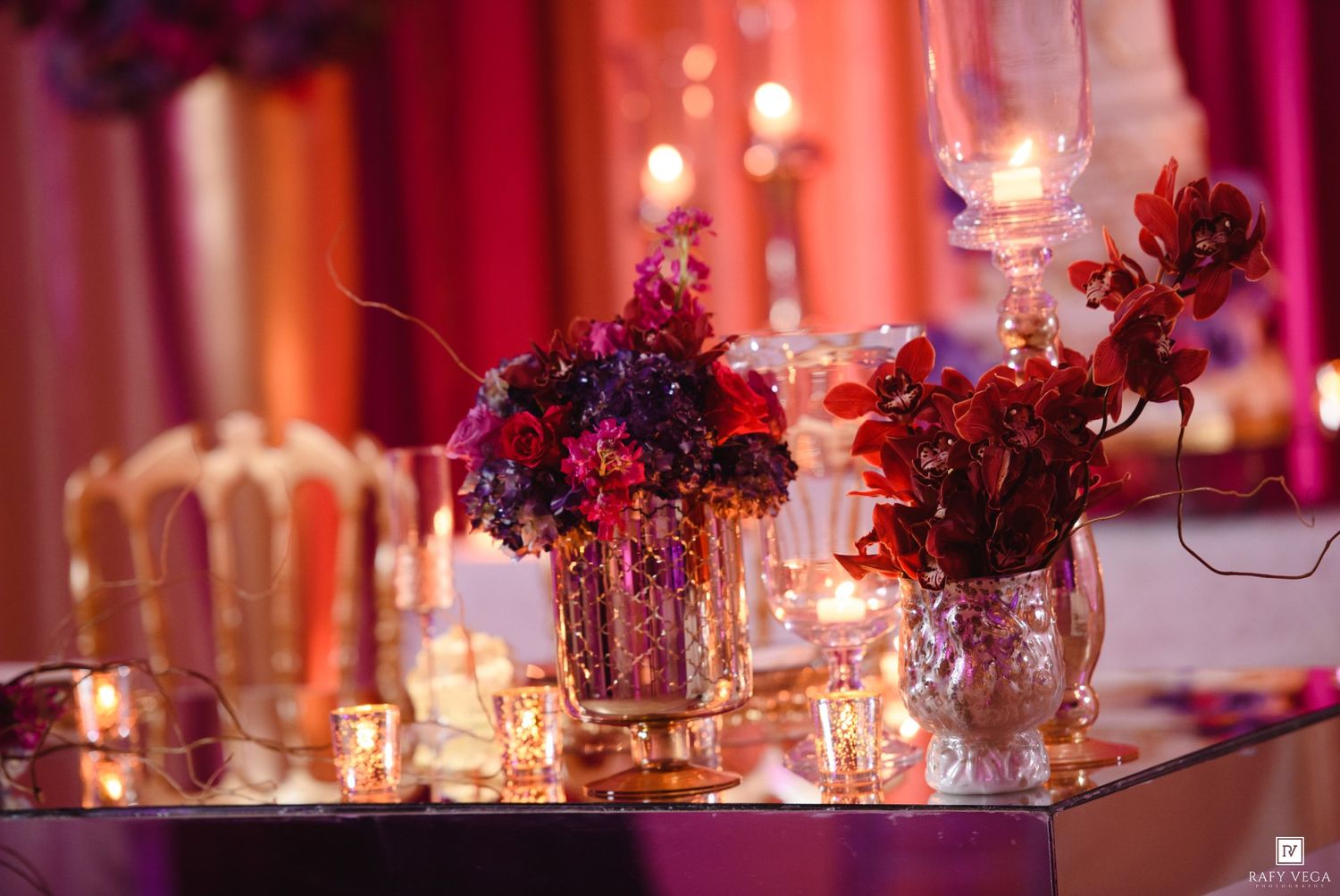 Elegant wedding reception table with red and purple floral arrangements in silver vases, candles, and warm pink lighting.