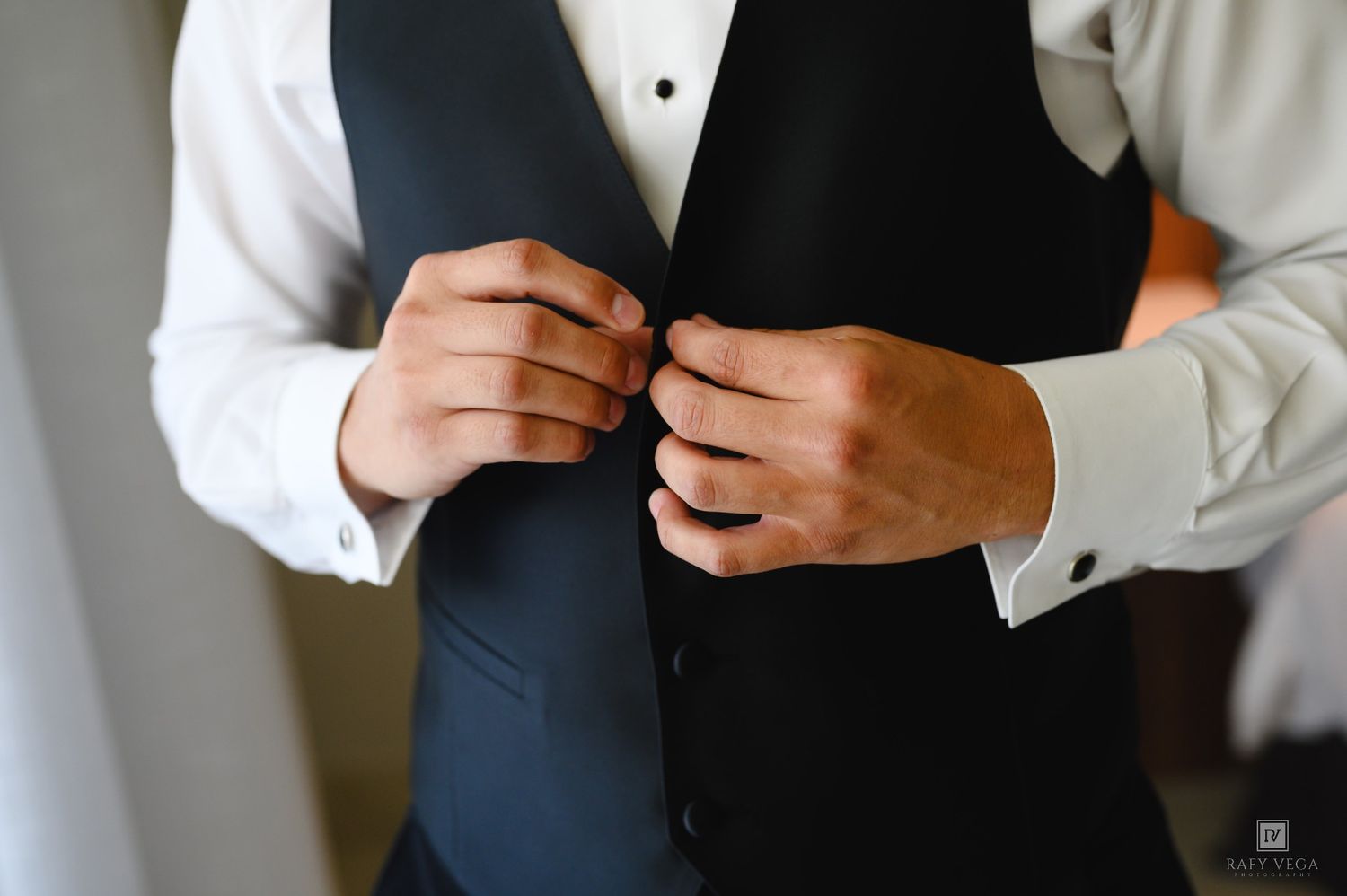 Groom adjusting his black tuxedo vest over a white dress shirt with cufflinks before wedding ceremony.