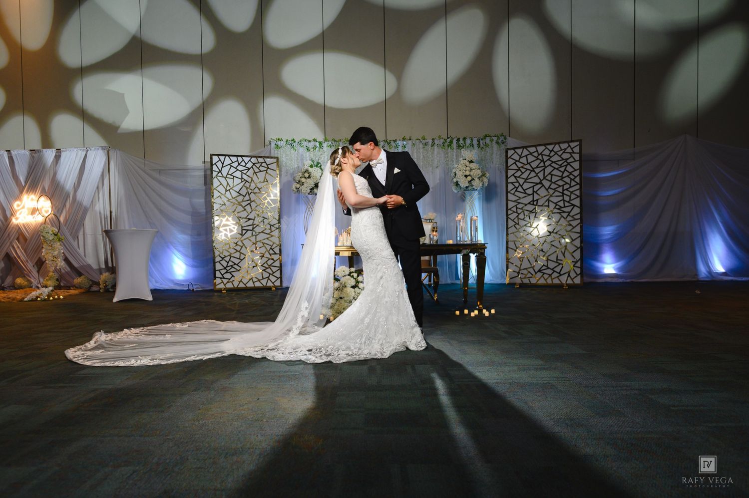 Bride and groom share first dance in elegant wedding venue with decorative lighting and floral backdrop.