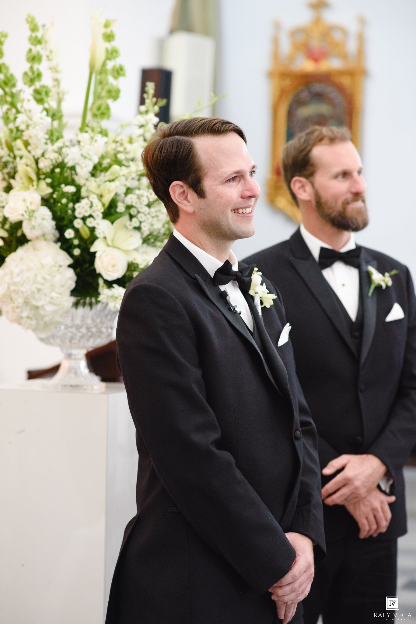 Two groomsmen in black tuxedos with white boutonnieres smile during a wedding ceremony in an elegant church.