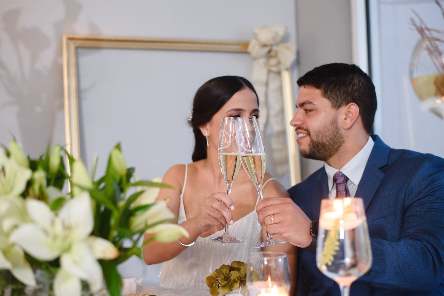 Couple clinking champagne glasses at wedding reception with white lilies and candlelight in background.