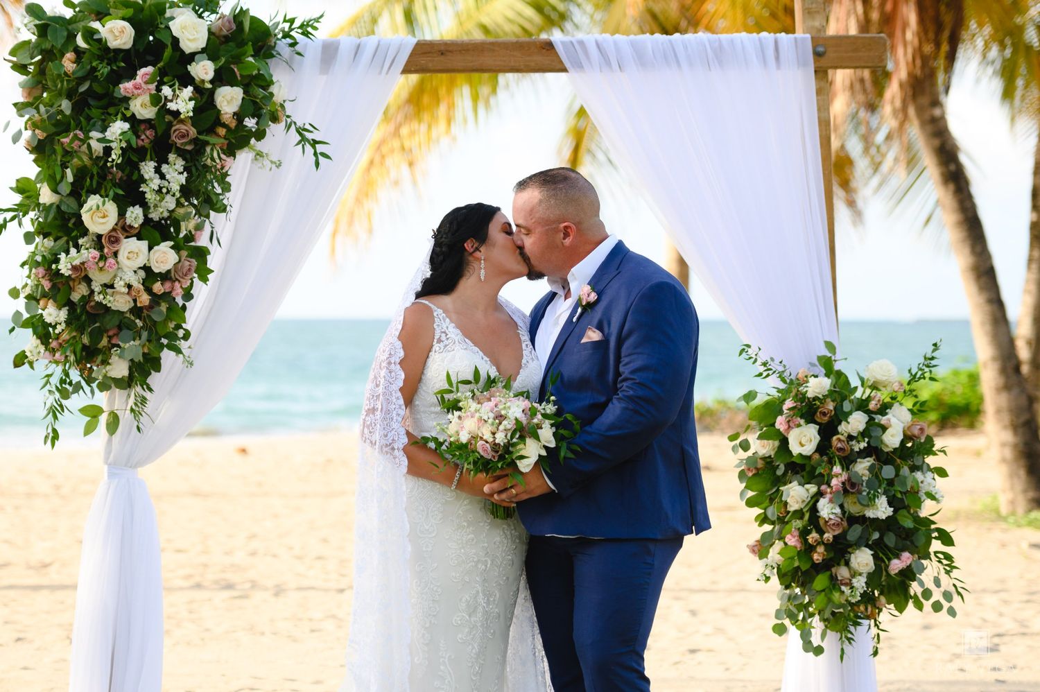 Bride and groom share first kiss at tropical beach wedding altar adorned with white drapes and blush floral arrangements.