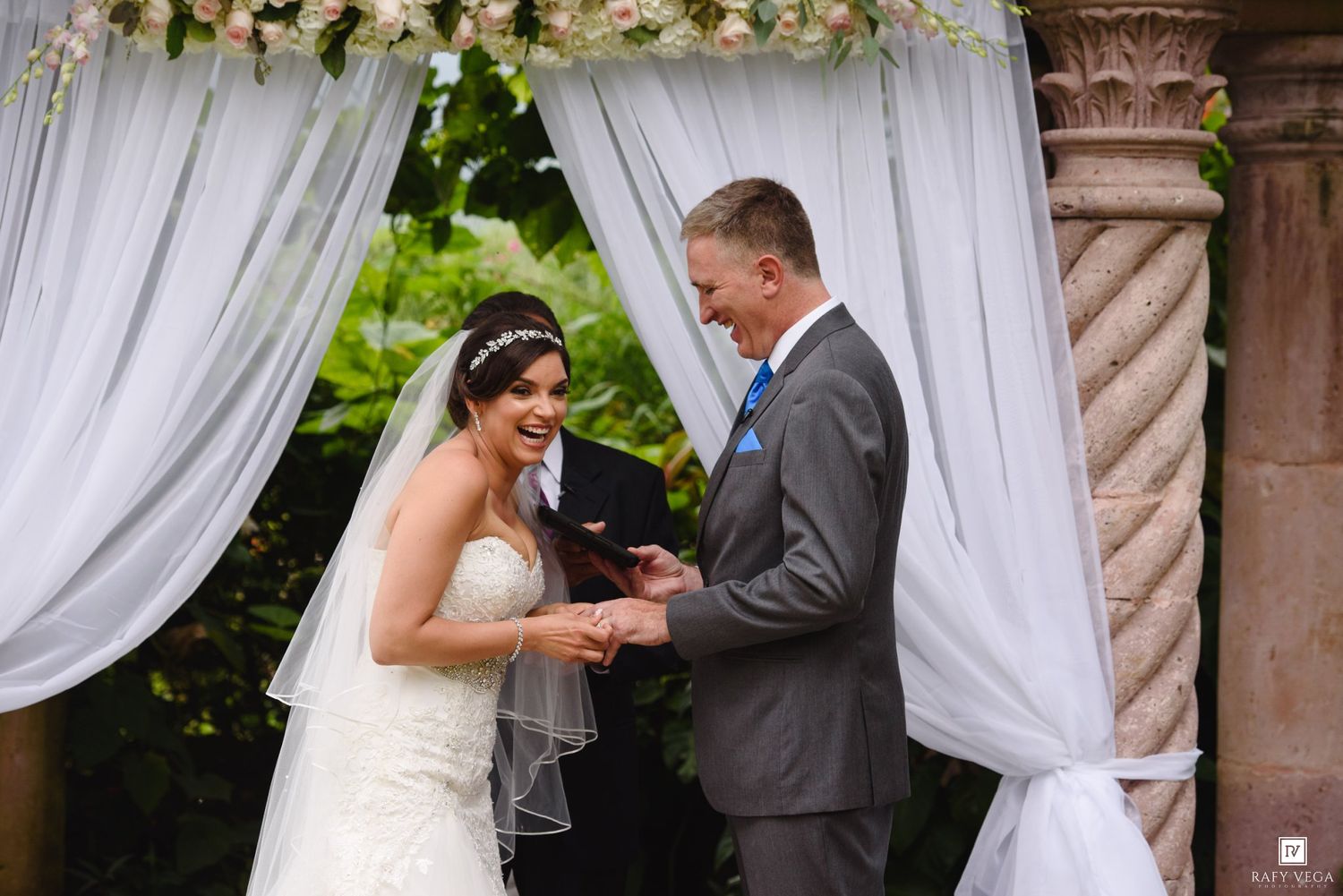 Bride and groom laugh joyfully during ring exchange under draped wedding arch with floral decor.