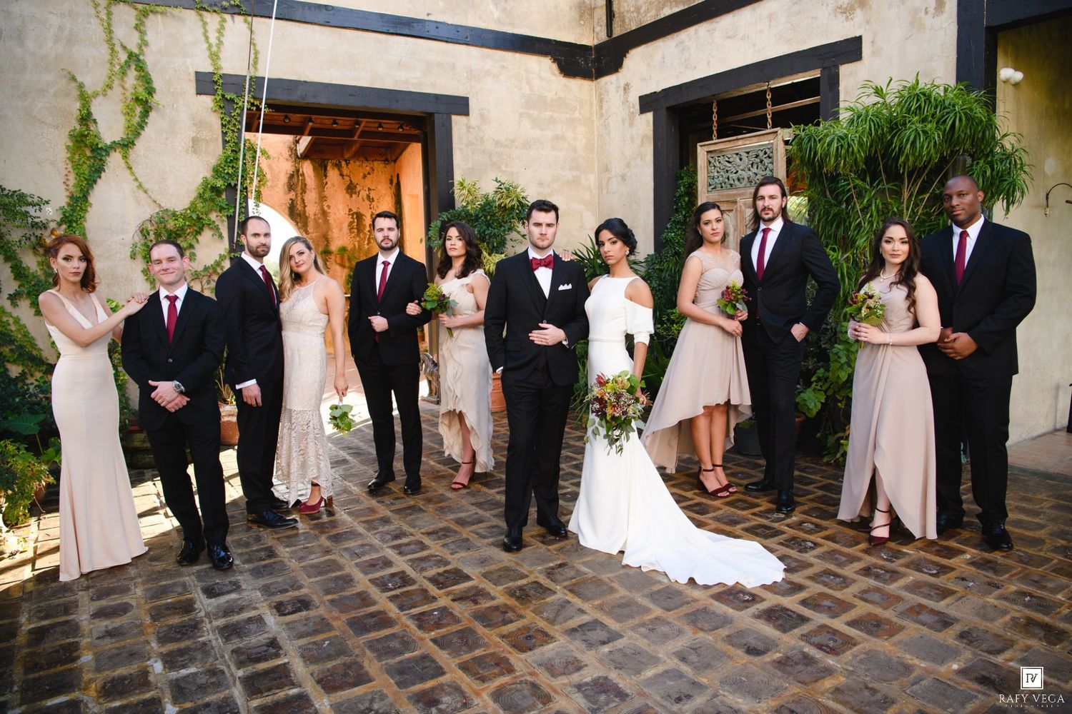 Wedding party poses in courtyard, bride in white gown flanked by bridesmaids in blush dresses and groomsmen in black suits.