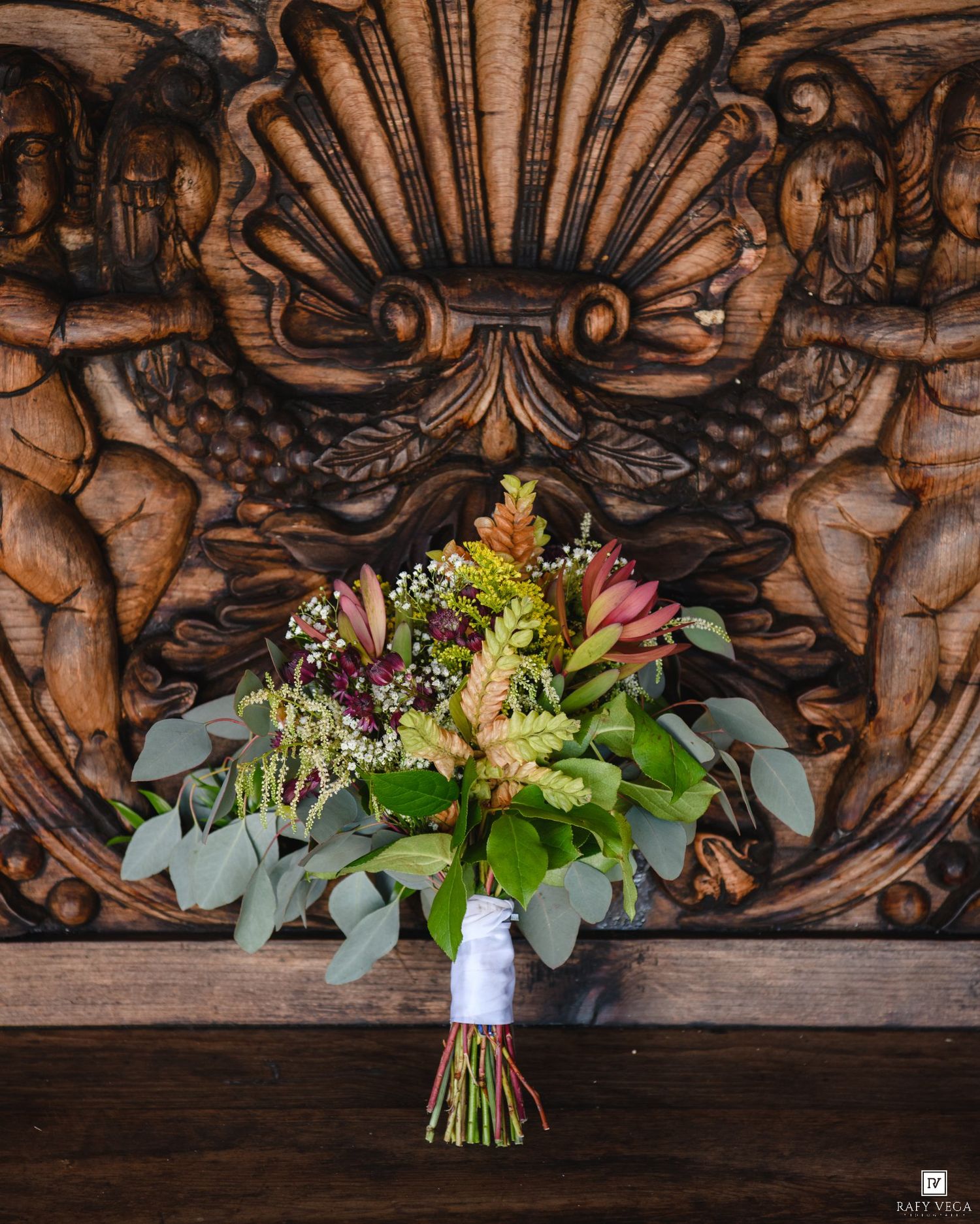 Bridal bouquet with eucalyptus, protea, and wildflowers against ornate carved wooden backdrop.