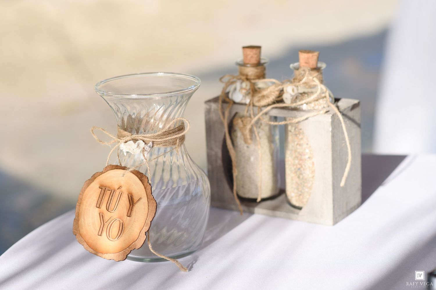 Rustic wedding sand ceremony vases with cork bottles, twine, and wooden tag reading Tu y Yo on white surface.