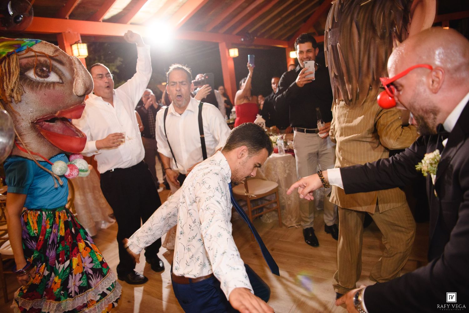 Guests dancing at a lively wedding reception with colorful puppets and red clown noses.