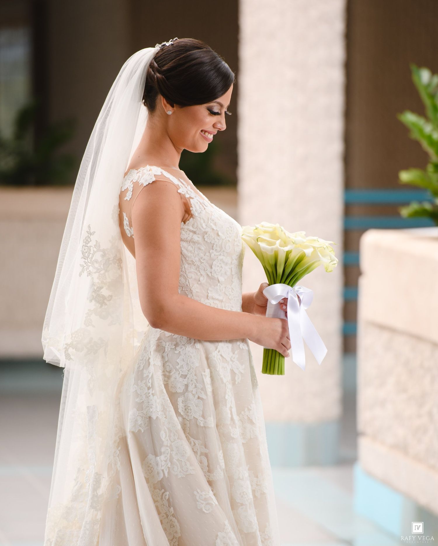 Bride in lace wedding gown and veil holding white calla lily bouquet with ribbon, smiling downward outdoors.