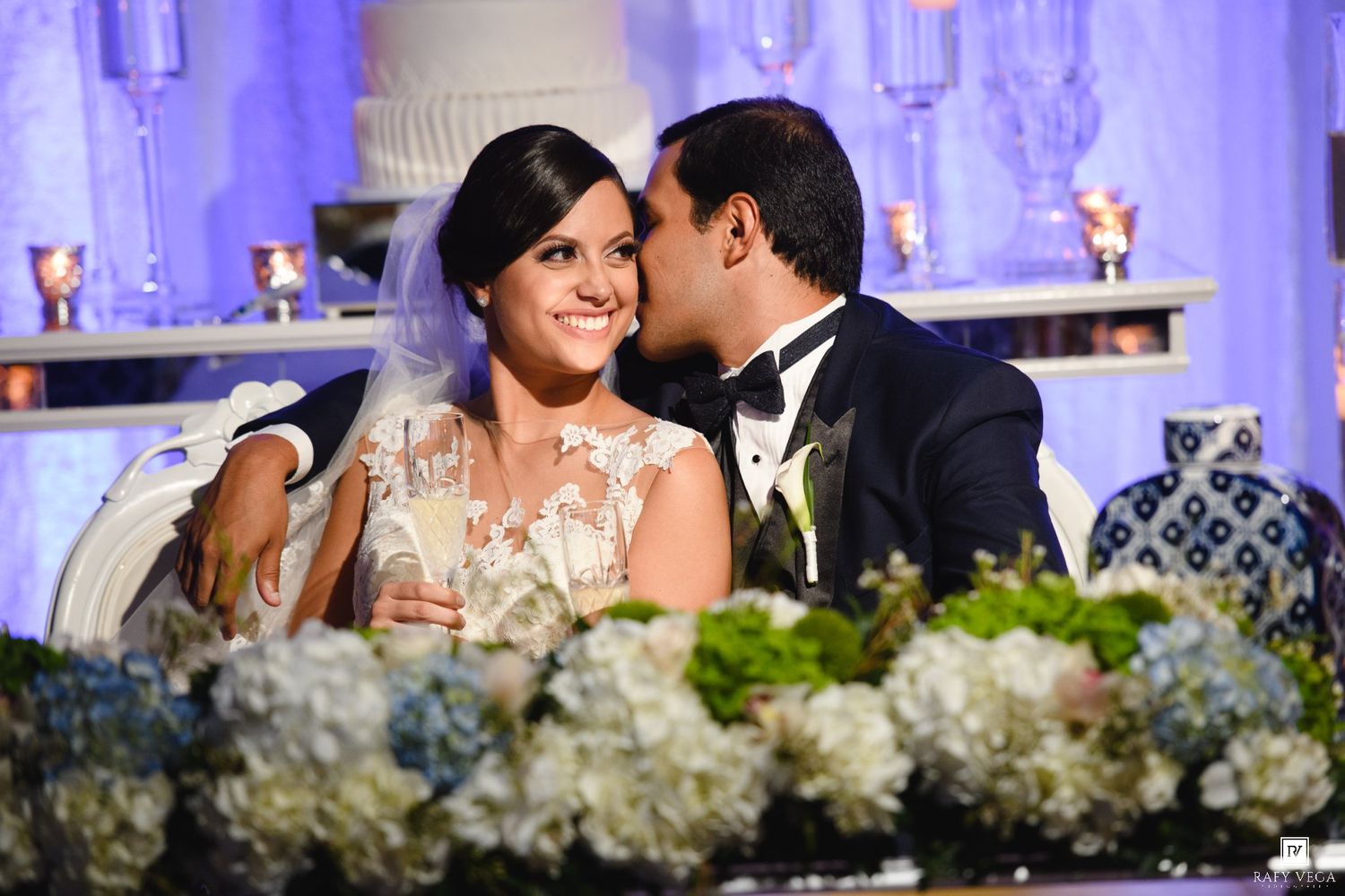 Bride and groom share a romantic moment at wedding reception sweetheart table with white hydrangeas and cake backdrop.