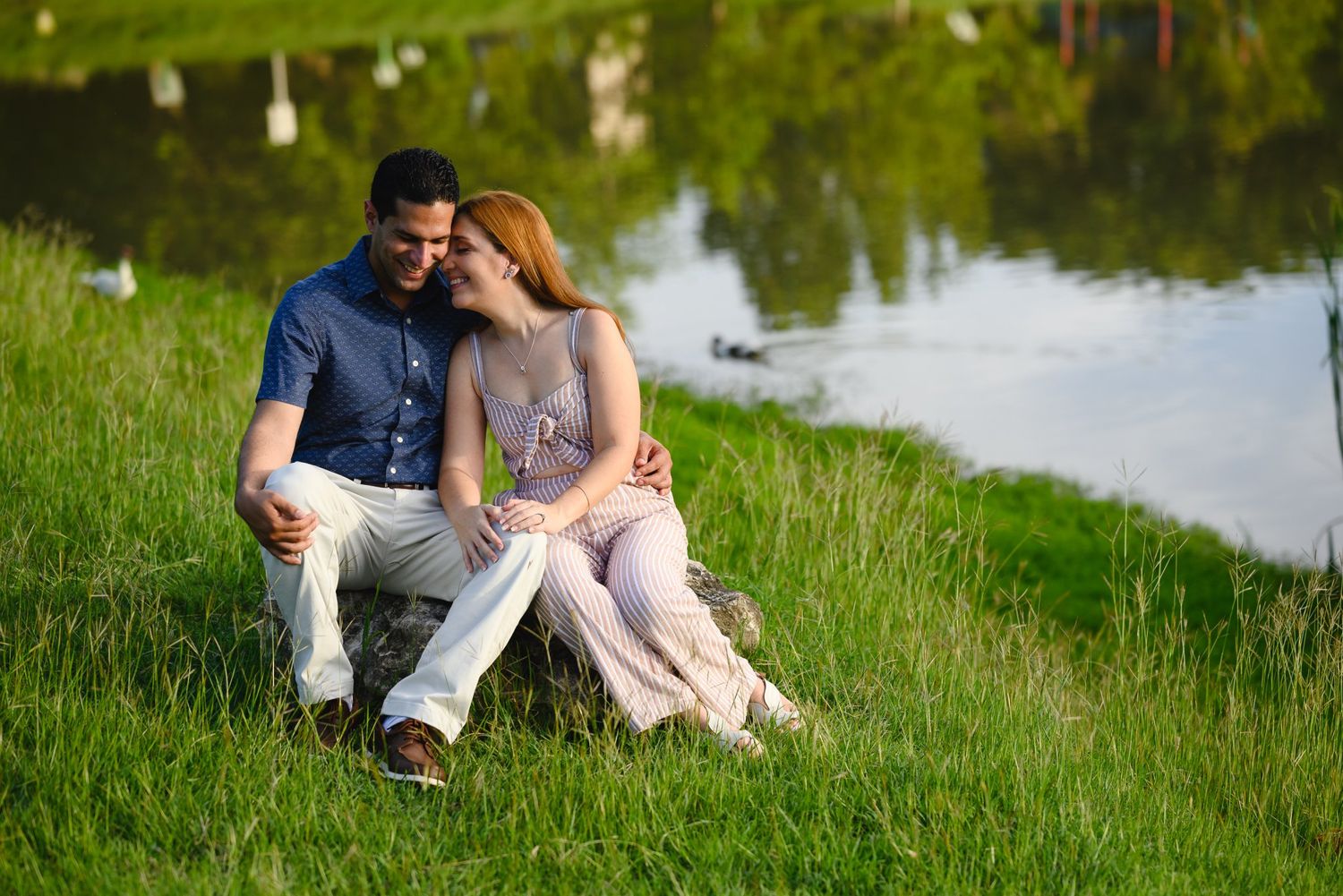 Romantic couple sitting together on grass by a serene pond, sharing a tender moment in warm sunlight.