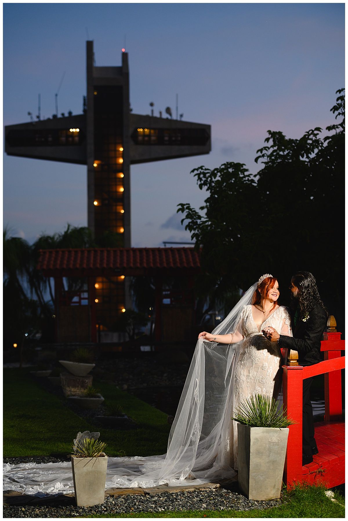 Bride in white gown and veil poses at dusk with illuminated church tower in background.