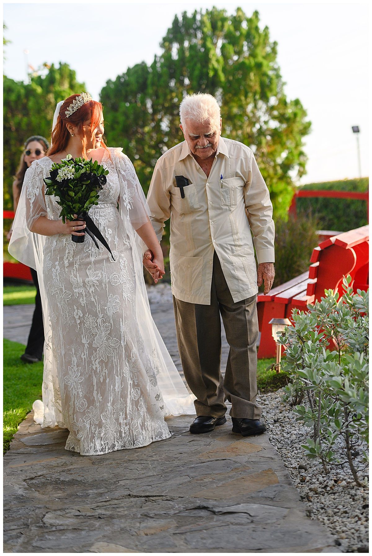 Bride in lace gown holding bouquet walks with elderly man on stone path at outdoor wedding venue.